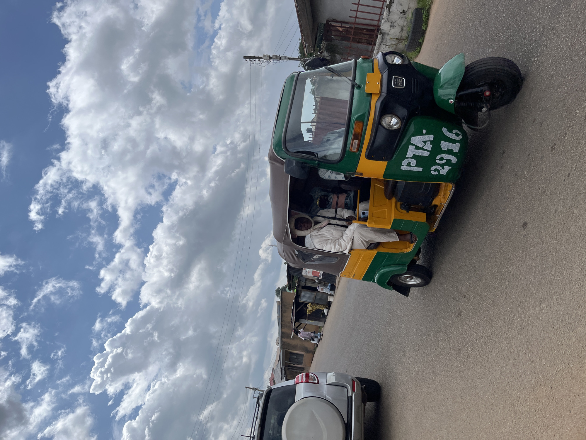 An outdoor, medium-shot, eye-level, natural light, photographic scene captures a street in the city of Jos, Nigeria, on a partly cloudy day. In the foreground, a green and yellow auto-rickshaw, known locally as a "Keke," drives on the right side of a paved road. The auto-rickshaw has a canopy, and a man in a white robe and head covering is visible in the driver's seat. The vehicle's license plate reads "PTA-2916." To the left of the auto-rickshaw, a silver SUV is parked, facing away from the viewer, with a spare tire mounted on its rear. In the background, several single-story buildings line the side of the road, some appearing to be shops or market stalls. A few people are visible walking near the buildings. The sky above is filled with a mix of blue sky and large, fluffy white clouds. Power lines are strung across the sky in the distance.