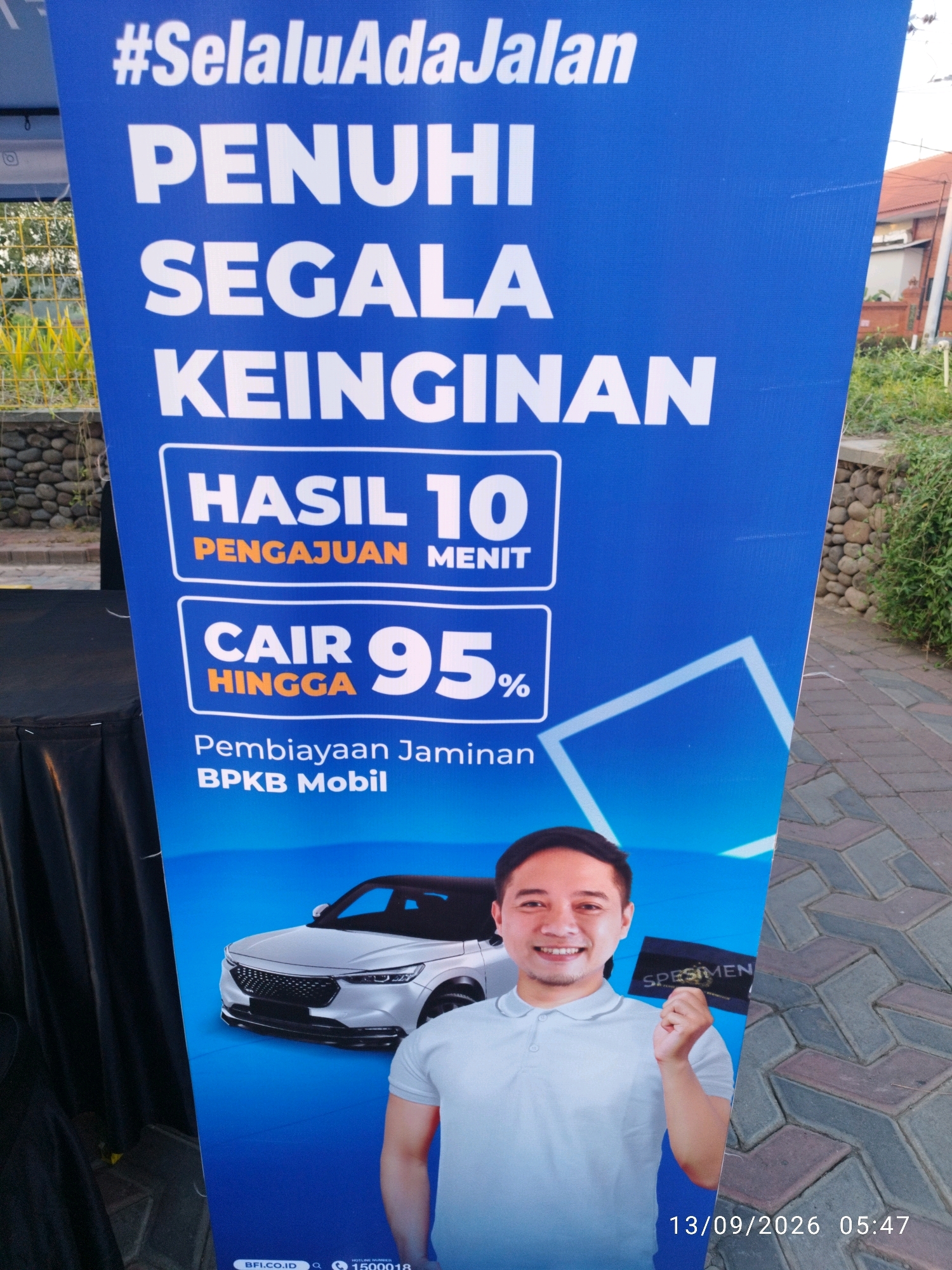 The image depicts a large blue banner with white and orange text, advertising financial services. The banner features the tagline "#SelaluAdaJalan PENUHI SEGALA KEINGINAN" which translates to "There's always a way to fulfill all desires". Below this, it states "HASIL 10 PENGAJUAN MENIT" (Results in 10 minutes for applications) and "CAIR HINGGA 95%" (Disbursement up to 95%). Further down, it mentions "Pembiayaan Jaminan BPKB Mobil" (Loan Guarantee with Car Vehicle Ownership Certificate).
A smiling man in a light blue polo shirt is shown on the right side of the banner, holding up a dark rectangular card labeled "SPESIMEN". A white car is depicted in the background on the left side of the banner. The bottom of the banner shows website information "BFI.CO.ID" and a hotline number "1500018". A timestamp at the bottom right reads "13/09/2026 05:47".
The banner is set outdoors, as indicated by the cobblestone pavement and greenery visible in the background. The overall scene appears to be a promotional display, possibly at a public event or along a street in Mojokerto, Indonesia, though the specific location within Mojokerto is not identifiable from the image alone. The lighting suggests it is daytime.
