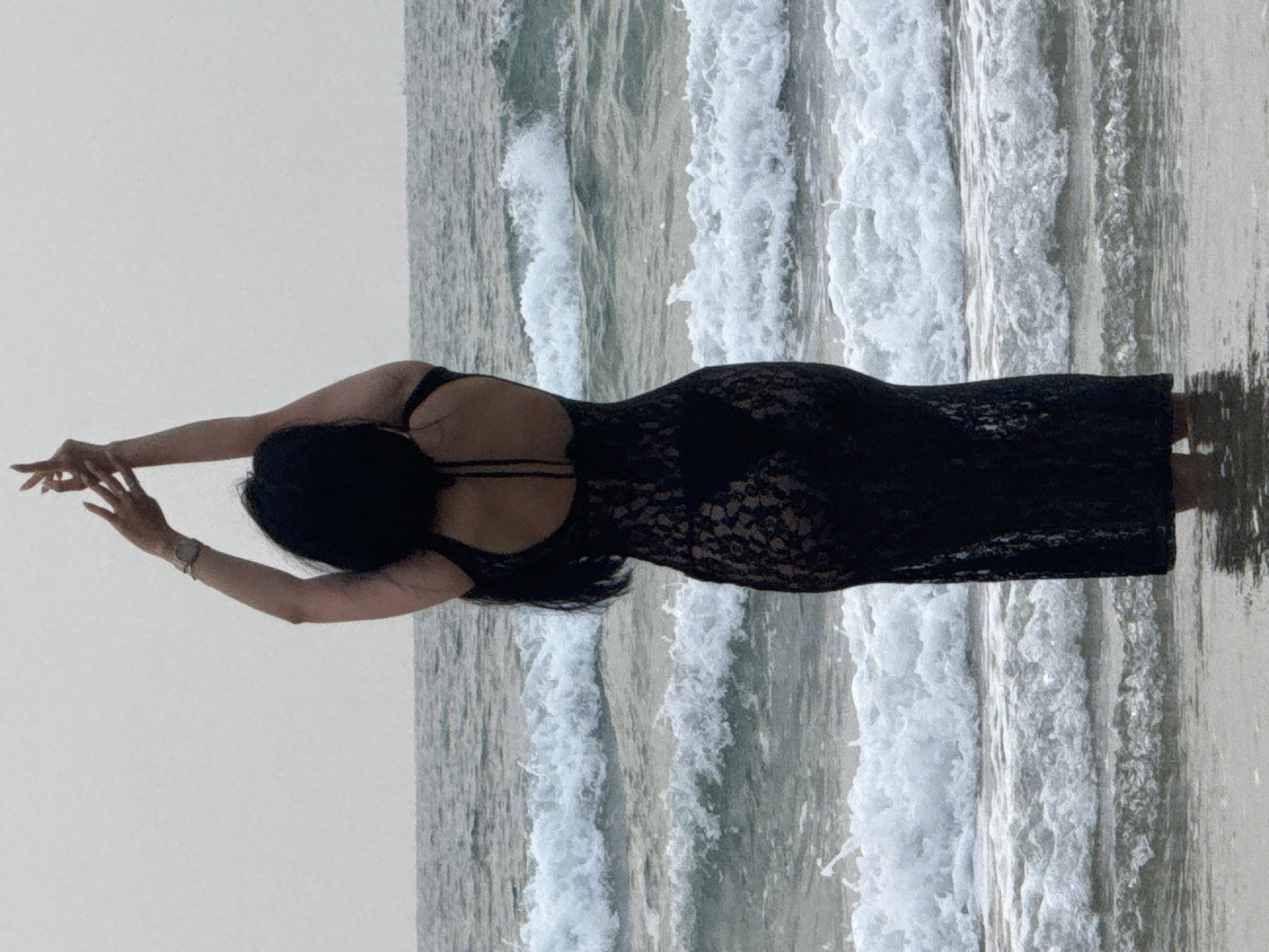 The image shows a woman in a black lace dress standing on a beach. Her arms are outstretched to either side. The ocean is in the background with crashing waves. The location is likely a beach in Đà Nẵng, Vietnam.