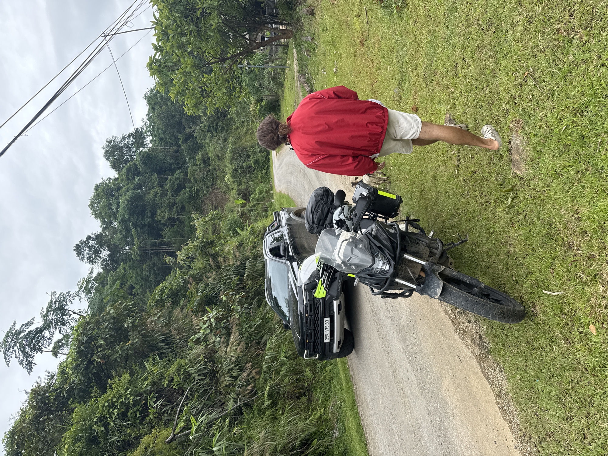 A person wearing a red jacket and light-colored shorts walks away from a motorcycle parked on the grassy shoulder of a rural dirt road. A black pickup truck is parked on the road in the background. The scene is surrounded by lush green vegetation and trees, suggesting a natural, possibly mountainous, environment. The sky is overcast. The person's hair is pulled back in a bun, and they are wearing sandals. The motorcycle is equipped with saddlebags. Two goats are visible near the person and motorcycle. The license plate of the pickup truck reads "29K-179.83". The overall atmosphere is that of a travel or adventure stop in a remote, green landscape. The location appears to be a road in Pù Luông Commune, Vietnam.