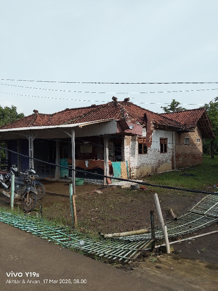 A view of a damaged brick house with a tiled roof, located in Kecamatan Anjatan, Indonesia, with coordinates of -6.389 latitude and 107.927 longitude. The house has a partially collapsed roof and a broken gate in front. There are two motorcycles parked in front of the house. The ground is covered in dirt and grass. Wires and cables are running across the sky.