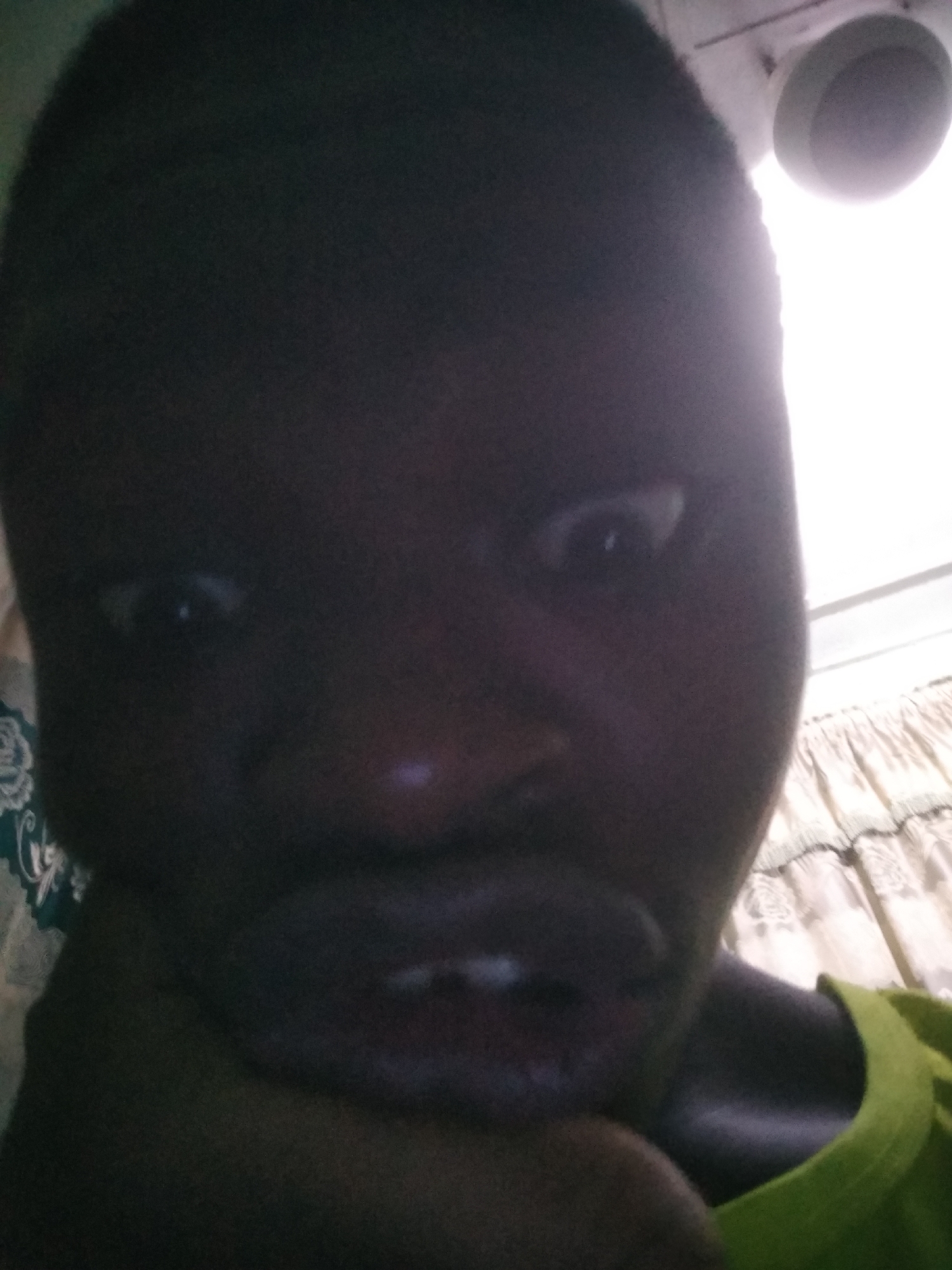 A close-up selfie of a person with dark skin. The person's face is the main subject, and their eyes are wide. The lighting is poor, making it difficult to discern detailed features. The person is wearing a light green shirt. The background shows a wall with patterned curtains. A ceiling light fixture is visible in the upper right corner. The location is unknown.