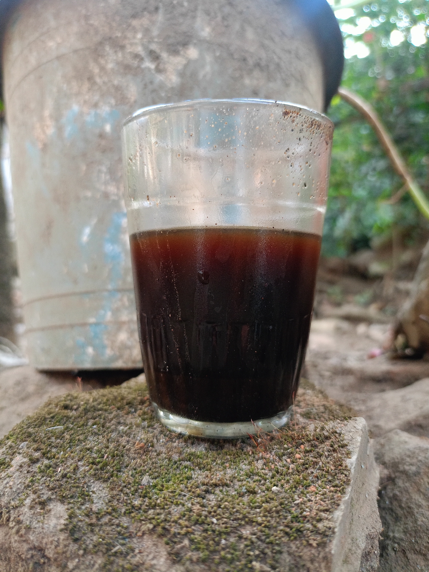 In Jelbuk, Indonesia, a close-up, eye-level shot focuses on a glass of dark liquid, likely coffee, resting on a moss-covered rock. The glass, with water droplets and small particles adhering to its exterior, contains a rich, opaque brown liquid that nearly fills it. In the background, a large, weathered, blue-and-gray plastic container looms, partially obscured by greenery. The overall environment suggests an outdoor setting, possibly a garden or yard, with a natural, rustic ambiance. The lighting is soft and diffused, indicating it may be overcast or the image was taken in shade. No people are visible in the frame.