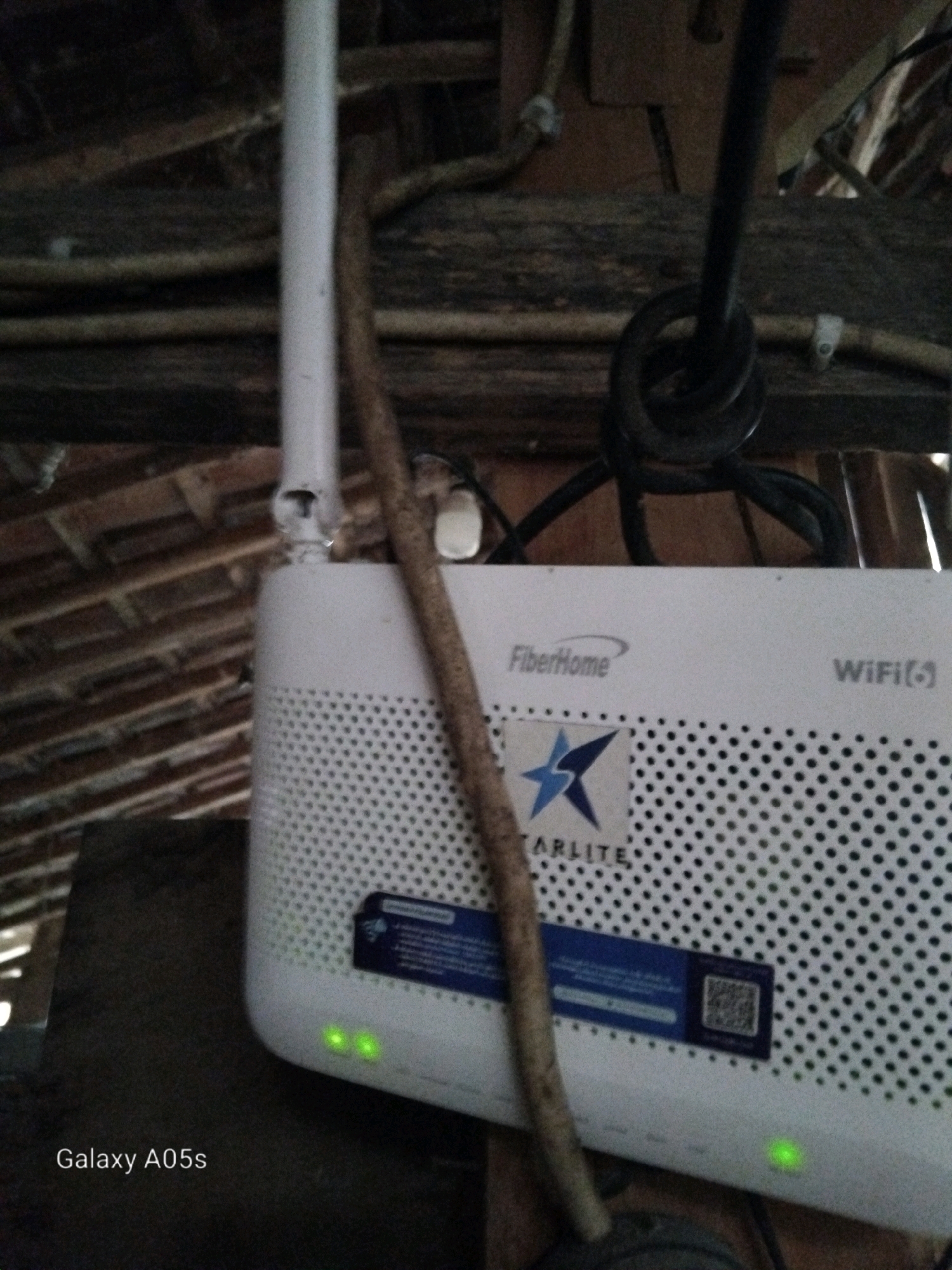 This is a medium shot, eye-level, close-up, interior photograph of a white wireless router, branded "FiberHome WiFi", with a "STARLITE" sticker on its side. The router is mounted on a dark surface, possibly a shelf or table, within what appears to be an attic or loft space, indicated by the visible wooden beams and roof structure in the background. The router has a vertical antenna and several green LED lights are illuminated on its front panel. A dark, coiled cable is visible above and behind the router. A thin, light-brown, stick-like object, possibly a twig or piece of wood, leans against the router, obscuring a portion of its front. The overall lighting is dim, suggesting natural light or minimal artificial illumination. The text "Galaxy A05s" is visible in the bottom left corner, indicating the device used to capture the image. The image is set in Karangayung, Indonesia.