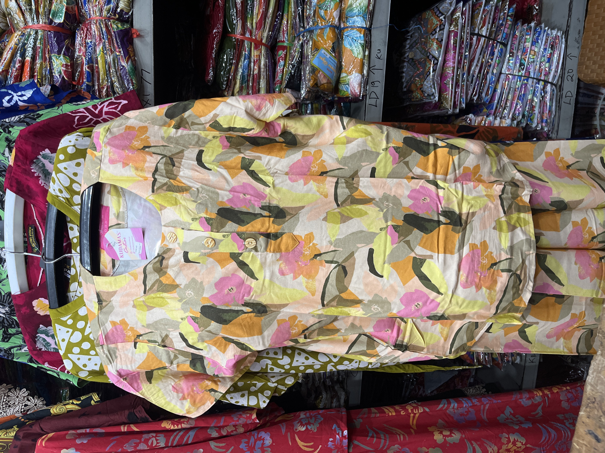 The image depicts a clothing store in West Jakarta, Indonesia. The foreground features a light-colored, floral-patterned shirt and matching pants on display, hanging on a rack. The shirt has a round neckline with two visible buttons and a tag that reads "KUDAMAS Couture". Behind the displayed garments, shelves are stacked with folded fabrics and clothing items, many of which are wrapped in plastic and secured with red bands. The overall environment suggests a retail setting focused on textiles and apparel.