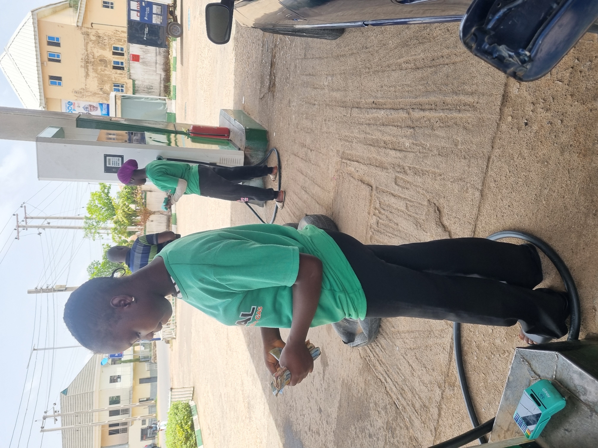 Here is a description of the image:
**Location:** Jos, Nigeria. The image depicts the scene at a gas station.
**People:** Two individuals, presumably gas station attendants, are visible. One is in the foreground, wearing a green t-shirt and black trousers, holding a wad of cash. The other is further back near a gas pump, wearing a green shirt and a purple head covering.
**Subjects/Objects:** A blue car is partially visible in the upper right corner. Gas pumps, hoses, a tire, a card reader (OPay branded) and buildings are also present.
**Actions:** The attendant in the foreground seems to be counting money. The other attendant is likely filling a vehicle with gas.
**Interactions:** The image implies a business transaction is taking place between the attendants and a customer filling their car with gas.
**Environment:** The gas station appears to be located in an urban area. The ground is a light, sandy color, possibly indicating a gravel or sandy surface. Buildings are visible in the background.
