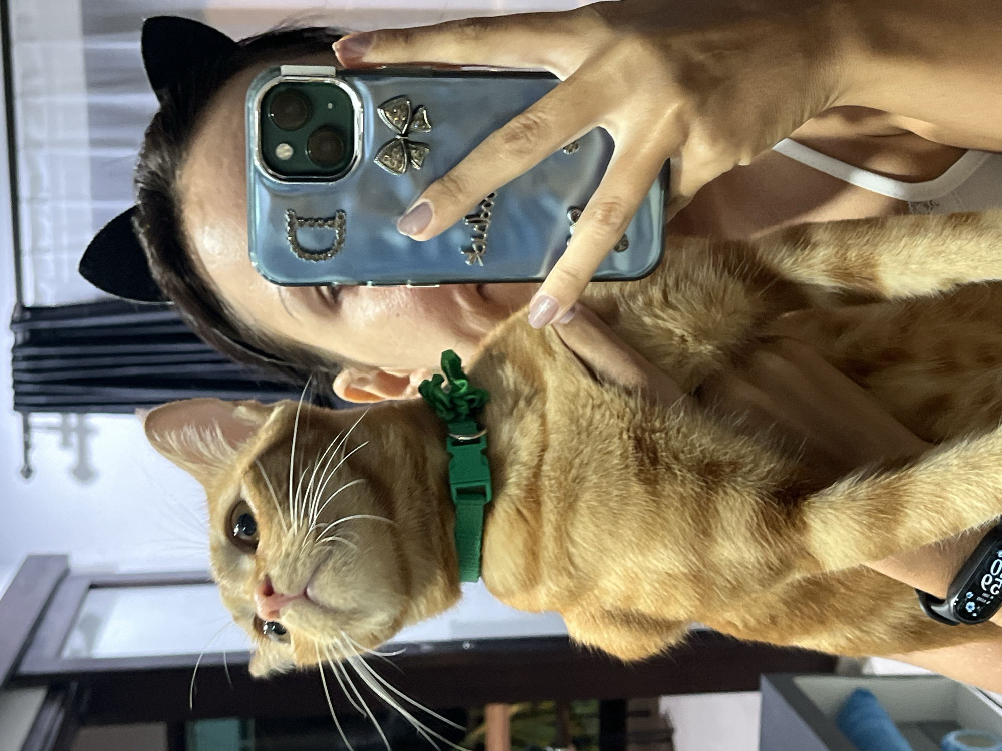 A woman is taking a selfie with an orange tabby cat. She is wearing black cat ears and holding a cell phone with a light blue case decorated with silver accents. The cat is wearing a green collar with a decorative green tie. The woman is wearing a smart watch on her left wrist. The background appears to be a room with a window. The location is Kuta Selatan, Indonesia.