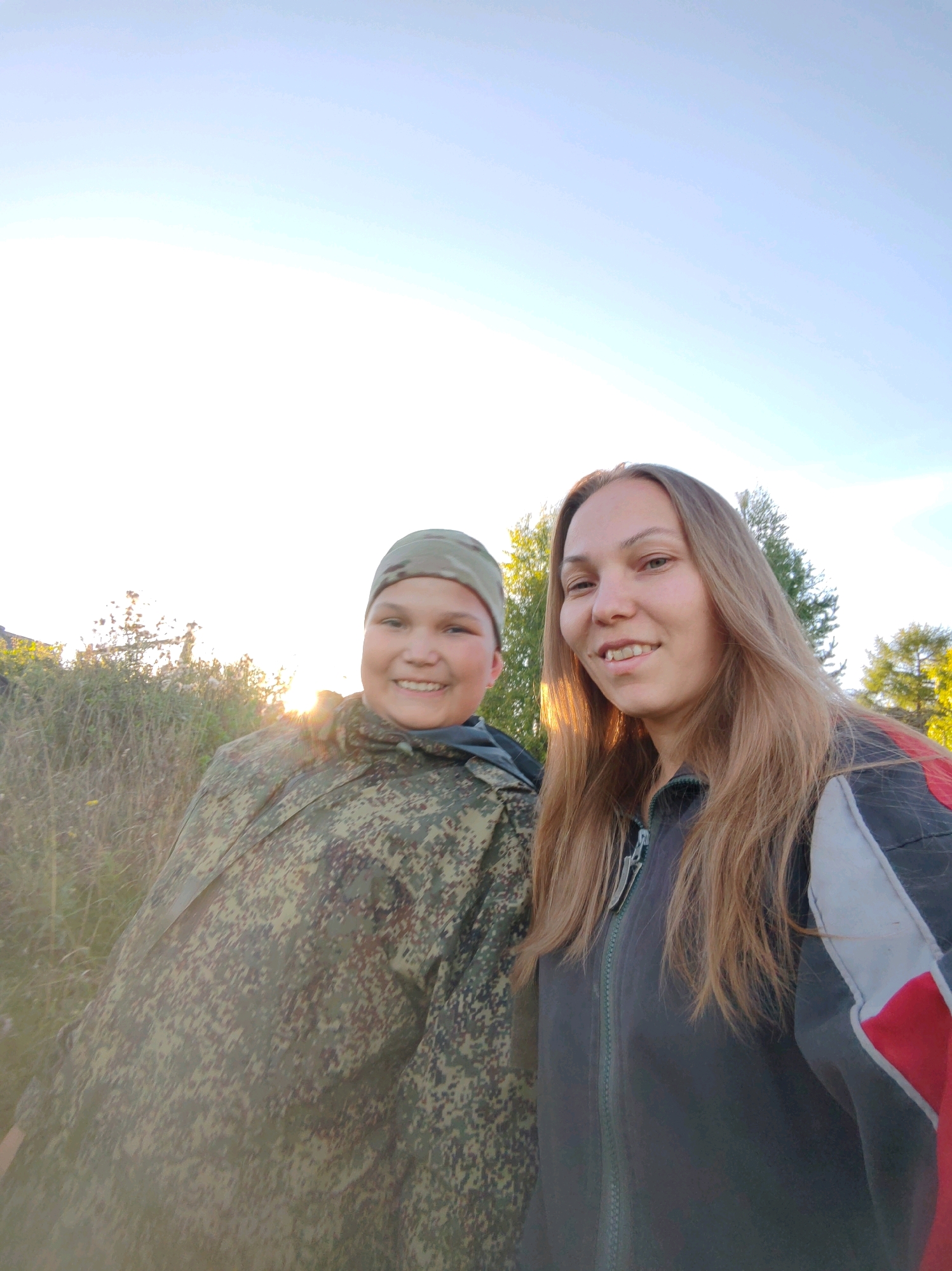 Two individuals, a child and an adult woman, pose for a selfie outdoors in what appears to be a rural or natural setting. The child, on the left, is wearing a camouflage hat and jacket, and is smiling broadly. The adult woman, on the right, has long, light brown hair and is wearing a dark jacket with red and white accents. The background consists of dry grass, some vegetation, and trees, with a bright, overcast sky. The lighting suggests it is either early morning or late afternoon. The specific location in the city of Чухлэм, Russia, is not discernible from the visual information alone.
