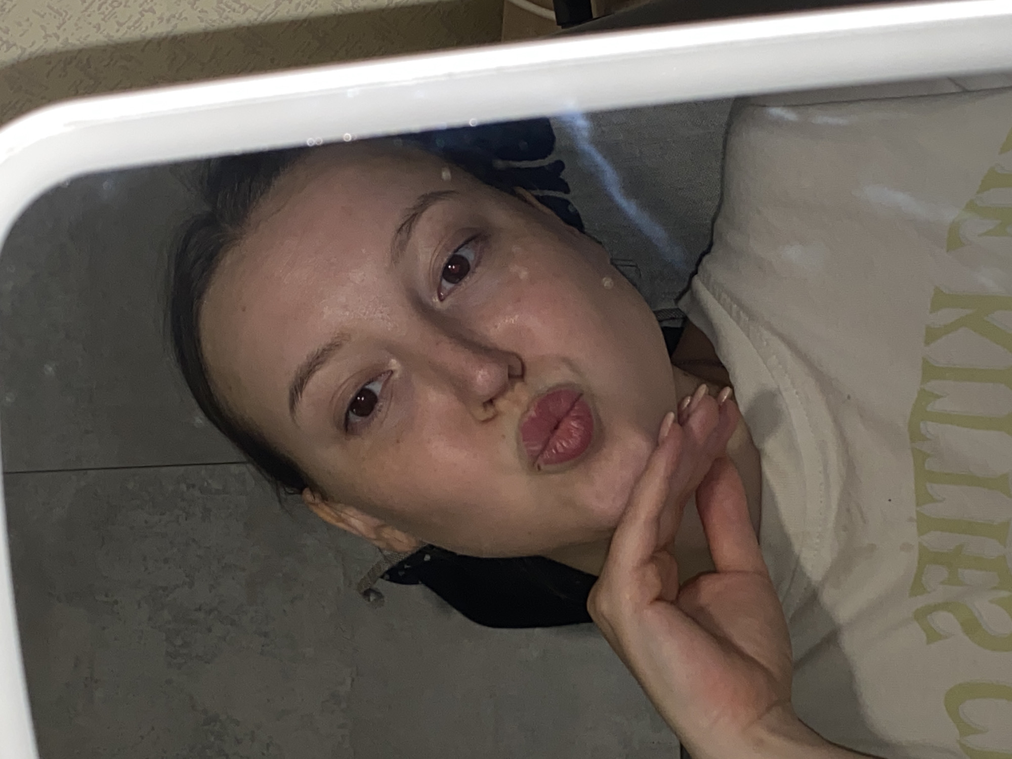 A person is reflected in a square mirror with rounded corners. The person, a fair-skinned individual, is looking at the reflection with pursed lips. Their right hand is raised near their face. They are wearing a light-colored t-shirt with writing on it. The reflection also shows a patterned bedspread in the background and light gray tiles on the wall. No further details about the location in Chaikovsky, Russia are discernible from the image.