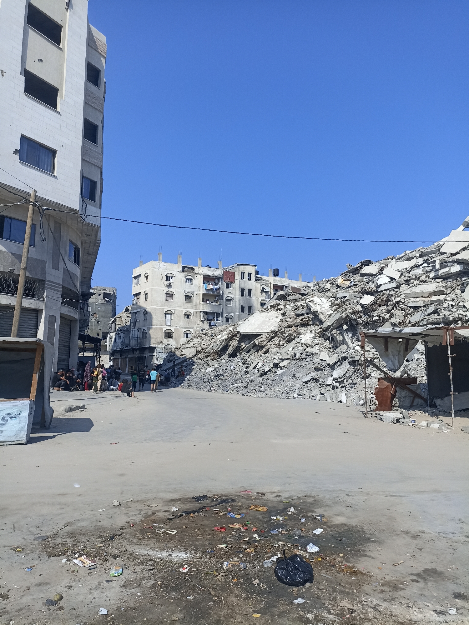 In the city of Gaza, Palestinian Territories, a street scene unfolds under a clear blue sky. On the left, a partially damaged, multi-story building stands, its exterior bearing signs of deterioration, with exposed concrete and dark window openings. Adjacent to it, a row of storefronts with closed metal shutters lines the street.
The center of the image is dominated by a vast pile of rubble, the remains of a destroyed building. This debris consists of concrete slabs, rebar, and other building materials, creating a rugged, uneven landscape. A makeshift structure, possibly a shelter or market stall made of corrugated metal and fabric, is partially visible at the base of the rubble pile on the right.
Several people are present in the scene. In the mid-ground, a group of individuals, some seated and others standing, are gathered on the street. Further down the street, other figures are visible, including a person in a blue t-shirt and shorts.
The ground in the foreground is a paved surface, stained and littered with debris. Scattered on the ground are various items, including wrappers, small pieces of plastic, and discarded food containers. A black plastic trash bag is also visible on the ground, near the debris. The overall environment suggests a scene of destruction and aftermath.