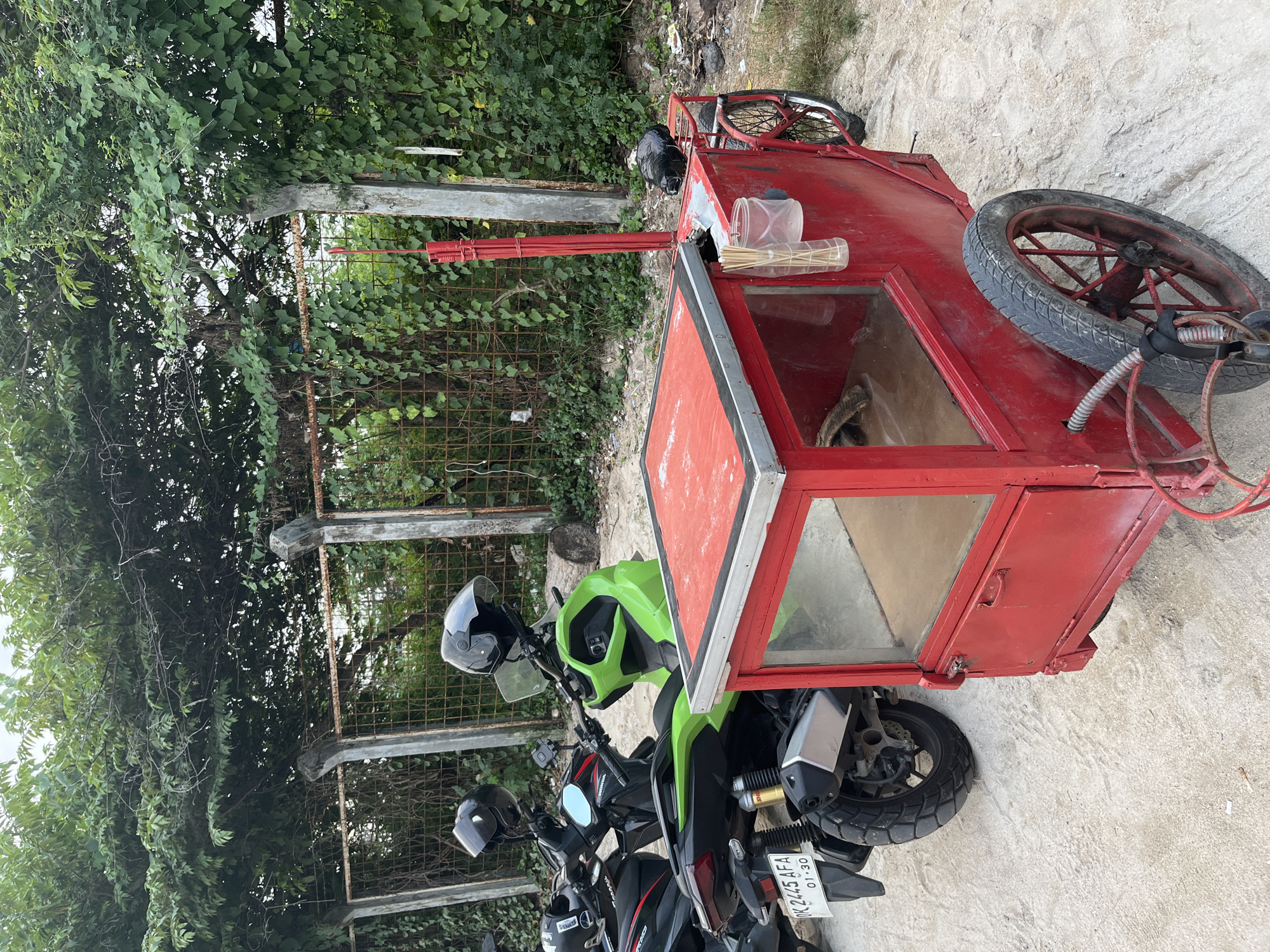 In Kuta, Indonesia, a red food cart is parked next to several motorcycles. The cart has a glass display case and a red canopy. Two helmets are visible on the motorcycles, one black and one white. The cart appears to be made of metal and has a single wheel in the front. The background is a fence covered in vines, with trees and foliage behind it. The ground is covered in sand and gravel.