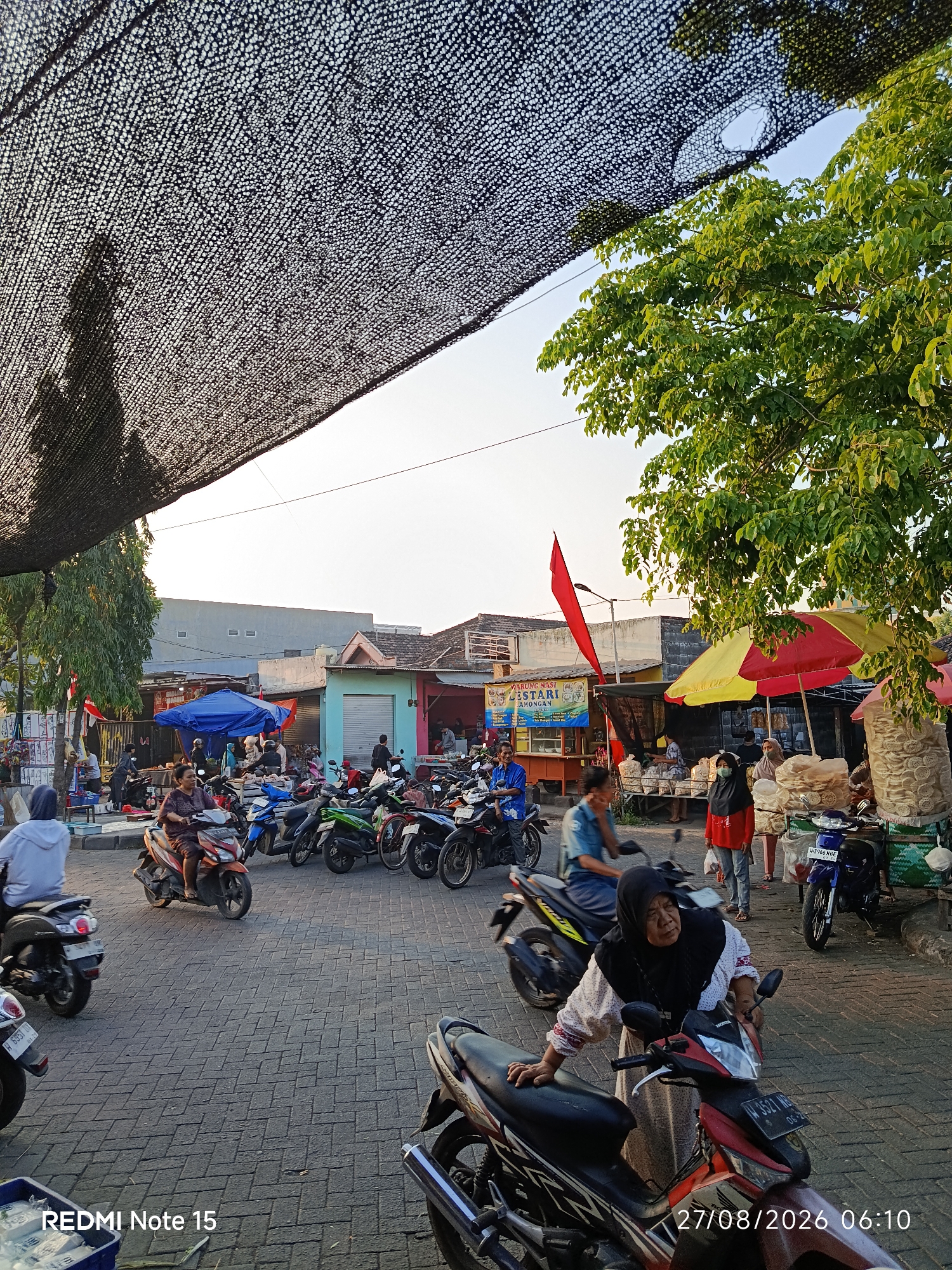 The image depicts a street scene in Keboansikep, Indonesia, likely a market area, under a large black mesh sunshade. Several people are present, some riding motorbikes, while others are standing or walking. Various stalls are set up, selling goods like food and snacks. Motorbikes are parked and in motion, indicating daily activity. The architecture of the buildings suggests a local Indonesian setting, with shops and houses lining the street. The lighting indicates it is daytime, possibly late afternoon given the angle of the sun and the long shadows.