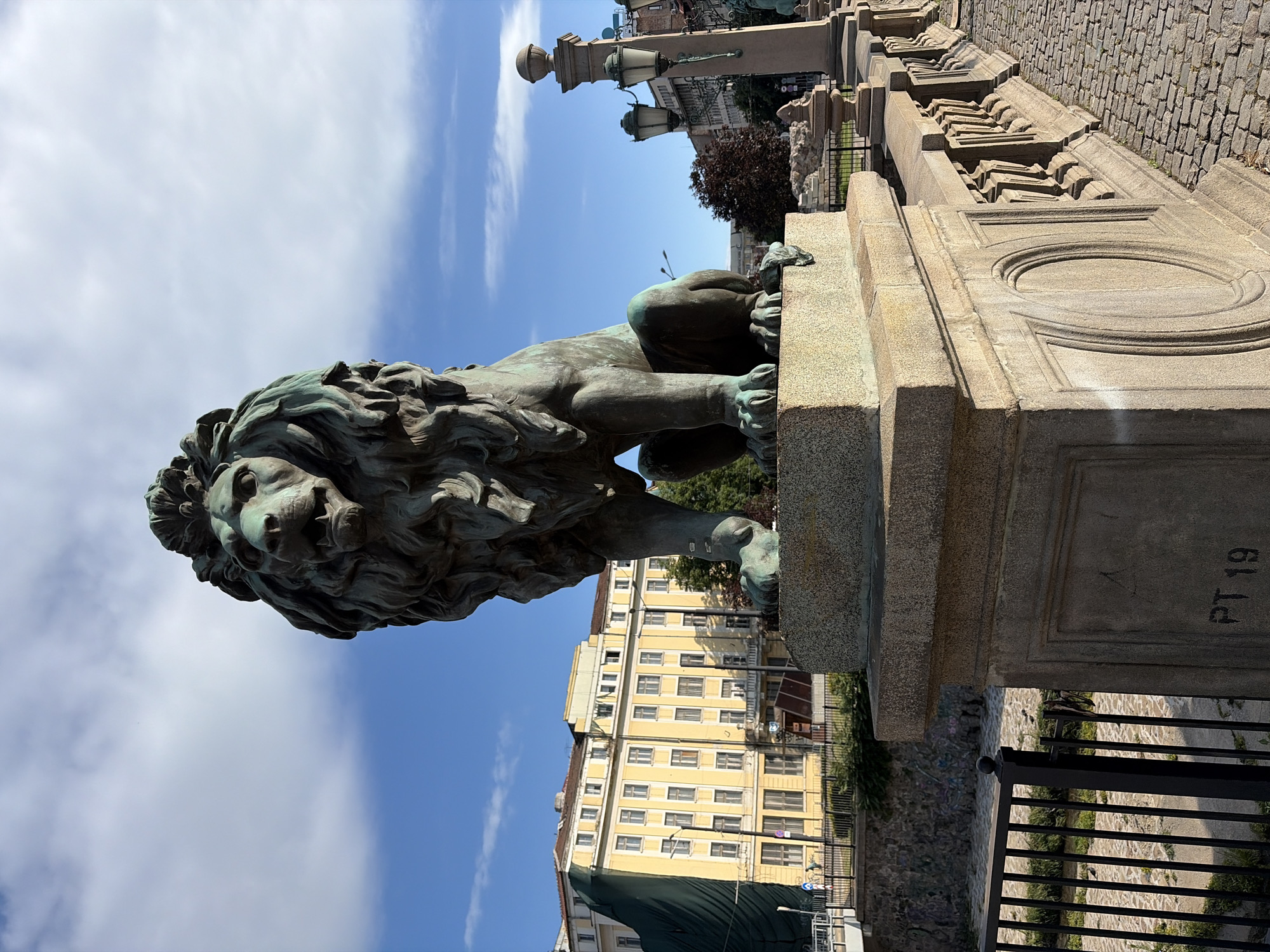 The image shows a bronze lion statue, a prominent landmark in Sofia, Bulgaria, specifically on the Eagles' Bridge (Orlov Most). The statue is positioned on a stone pedestal, with its gaze directed forward. Behind the statue, a large, yellow building with multiple windows stands, partially covered by a green tarp on the left side. The bridge itself is paved with cobblestones and features ornate stone railings with decorative elements. A black metal fence runs along the side of the bridge, overlooking a dry riverbed or canal with some vegetation and graffiti on the stone walls. Several lampposts, in a traditional style, are visible on the bridge. The sky above is blue with scattered white clouds. The scene is captured in daylight, suggesting it is outdoors.
