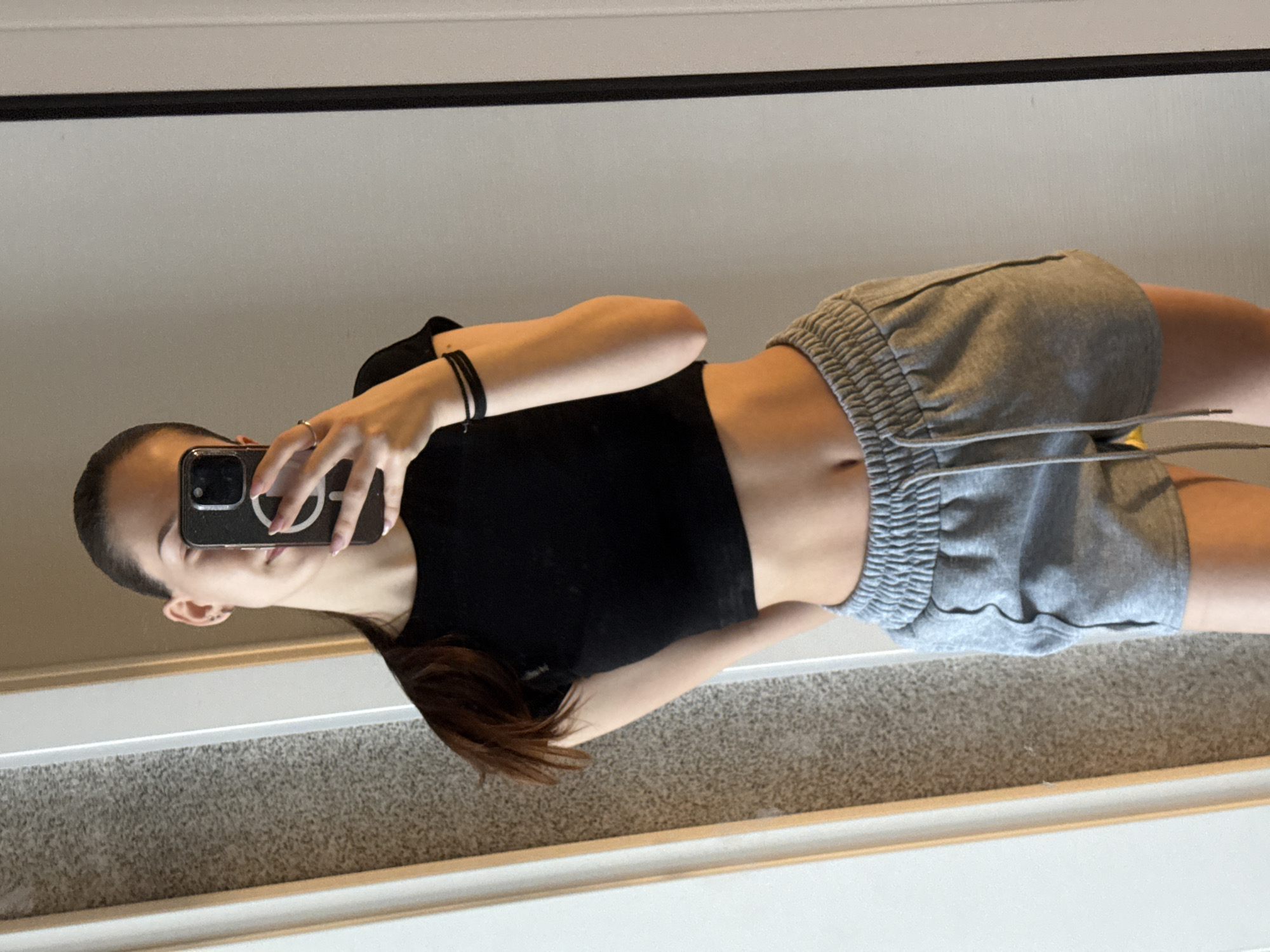 A young woman takes a selfie in a mirror. She is wearing a black crop top and gray sweat shorts. The background shows a bedroom with light carpet and white trim. The location is not discernible to be Sofia, Bulgaria without further information.