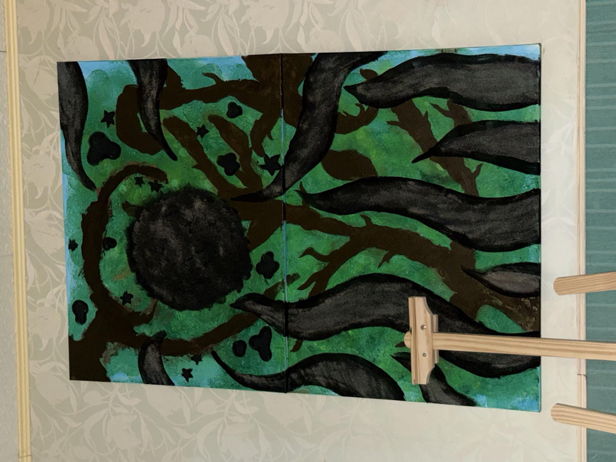 A framed abstract painting hangs on a light-patterned wall. The painting is predominantly green and blue with black and brown elements depicting shapes resembling leaves, branches, and a central dark circle. A light wood stretcher bar is attached to the right side of the framed painting. The environment is indoors. There is no indication of the location being Domont, France.