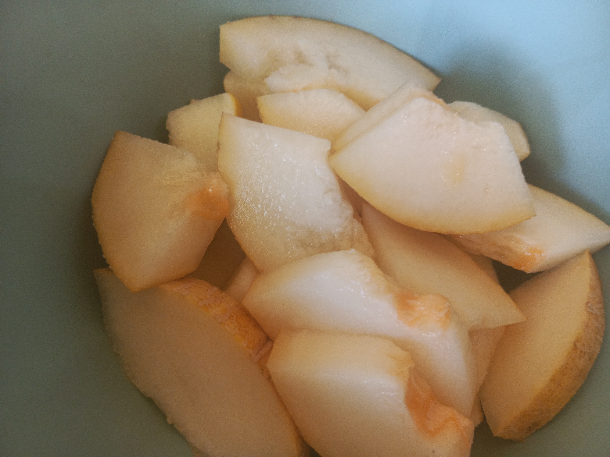 A close-up, overhead shot shows a pile of sliced cantaloupe in a light blue bowl. The cantaloupe is cut into wedges, with the rind still attached to some pieces. The flesh of the fruit is a pale orange-yellow color, and some pieces show the texture of the flesh with small seed cavities. The bowl appears to be made of a smooth, matte material. The lighting is bright, suggesting it may be daytime. The scene is simple and focuses entirely on the fruit. No people or other objects are visible. No text is present in the image. The context of Kolpino, Russia, is provided but not visually evident from the image itself.