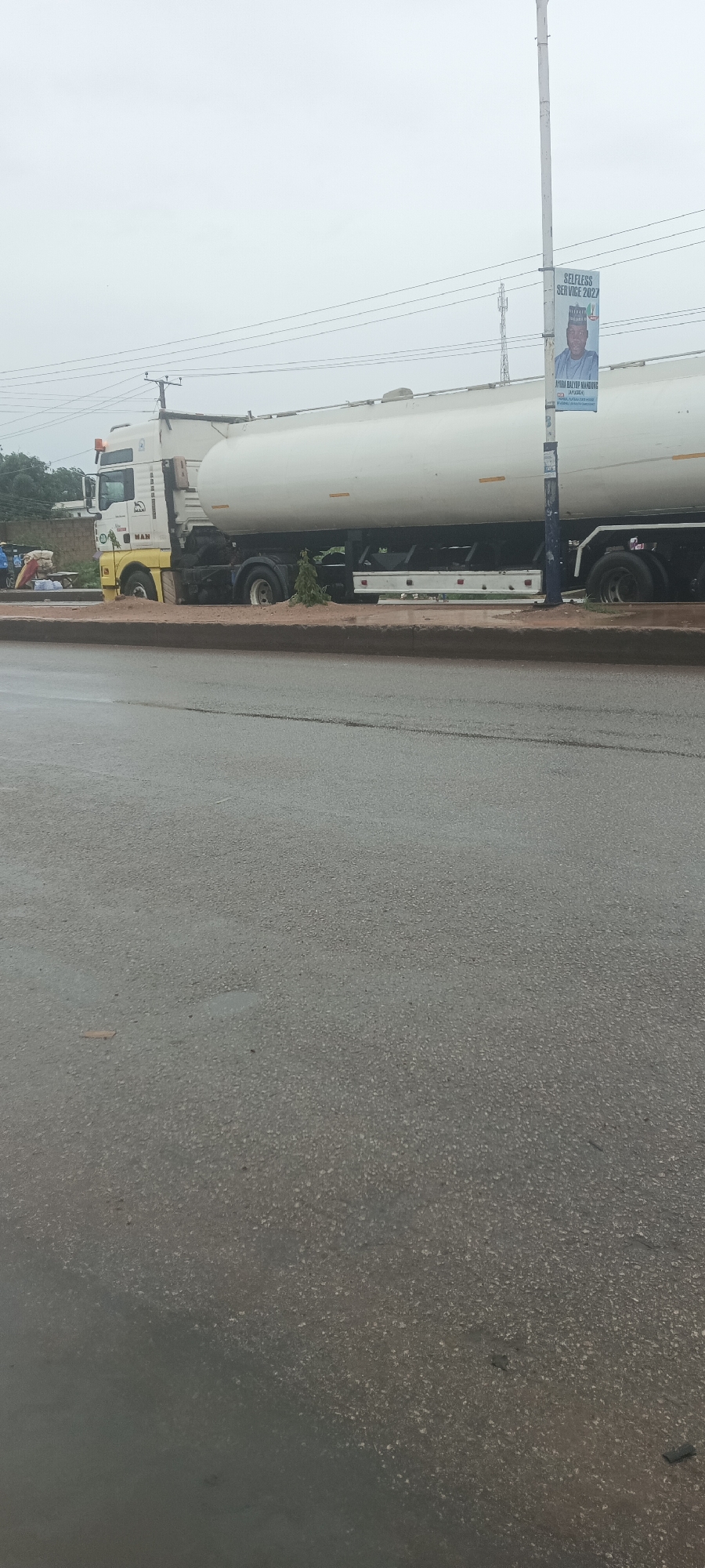 An outdoor, eye-level shot captures a large white tanker truck partially off a main road, parked or stuck on an unpaved median strip. The front wheels of the truck are resting on the sandy/earthy ground of the median, while the main body and rear wheels remain near the asphalt. The truck is a MAN brand vehicle, identifiable by the logo on its cabin.
The road surface in the foreground appears damp, suggesting recent rain or overcast weather. Above the truck, an overcast, light gray sky dominates the upper portion of the image.
To the right of the truck, a utility pole stands with overhead wires traversing the scene. A banner is affixed to this pole, featuring a man's portrait and text. Visible text on the banner includes "SELFLESS SERVICE 2027", "AYUBA DALIUP MANDUNG", "JP LEADER!", and "VOTE FOR", followed by a phone number (07038XXXXXX).
In the far left background, beyond the truck's cabin, a few indistinct individuals are visible, standing near what appears to be some objects or debris. The setting is consistent with a roadside in Bukuru, Nigeria, as provided by the context. The time of day appears to be daytime, under cloudy skies.