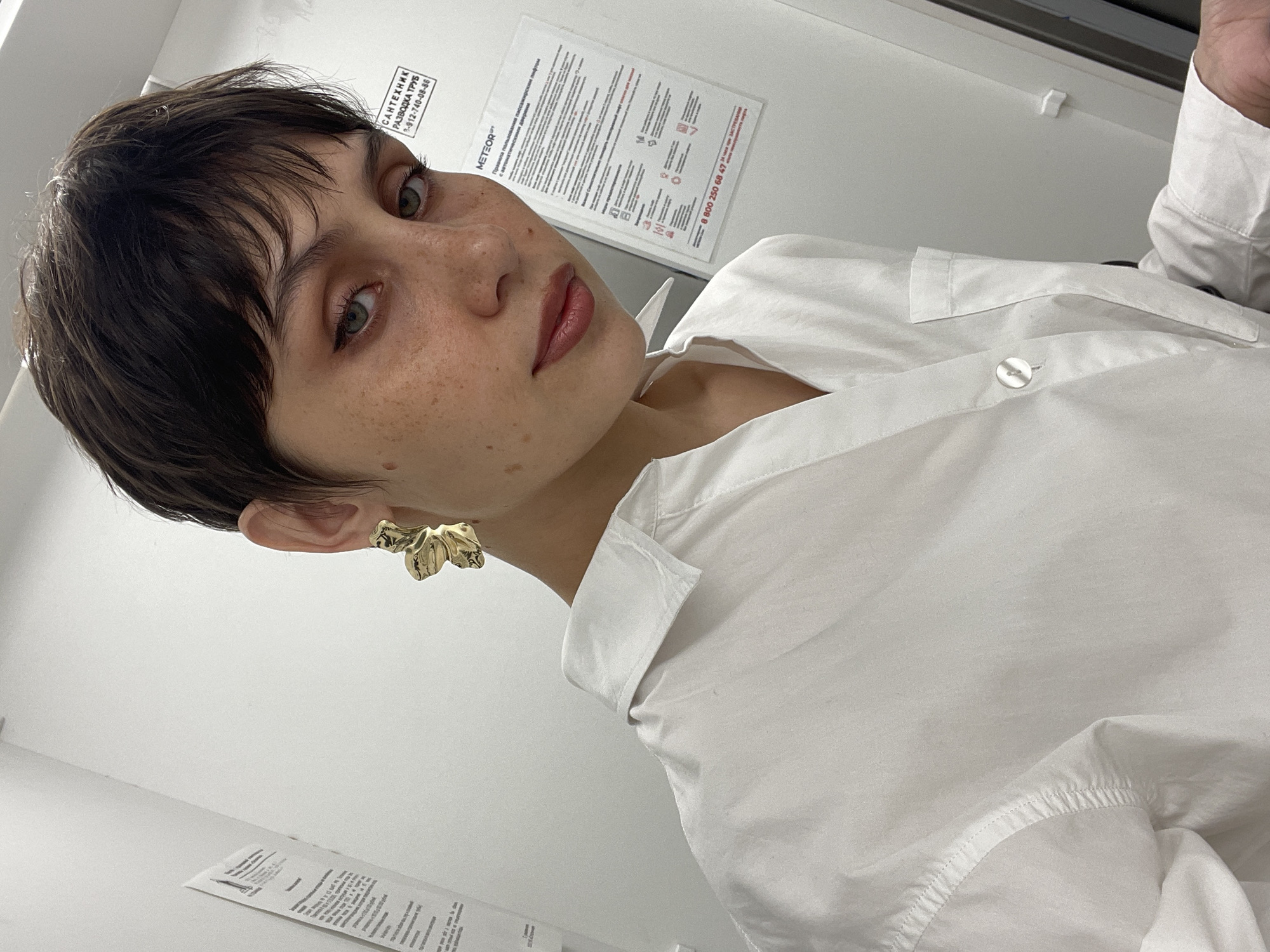 A close-up, low-angle shot depicts a young woman with short, dark brown hair and fair skin. She has visible freckles and is wearing a white button-down shirt. She is wearing a large, gold-colored earring in her left ear. The background consists of a white wall with several printed notices or papers affixed to it. One notice, partially visible, includes text that appears to be in Russian, and indicates the word "METEOR" and a phone number: "8 800 250 68 47." The notice also has the words САНТЕХНИК (Plumber) and РАЗВОДКА ТРУБ (Plumbing) with a phone number 9-912-740-08-86. The city of Izhevsk is not visible in this image.