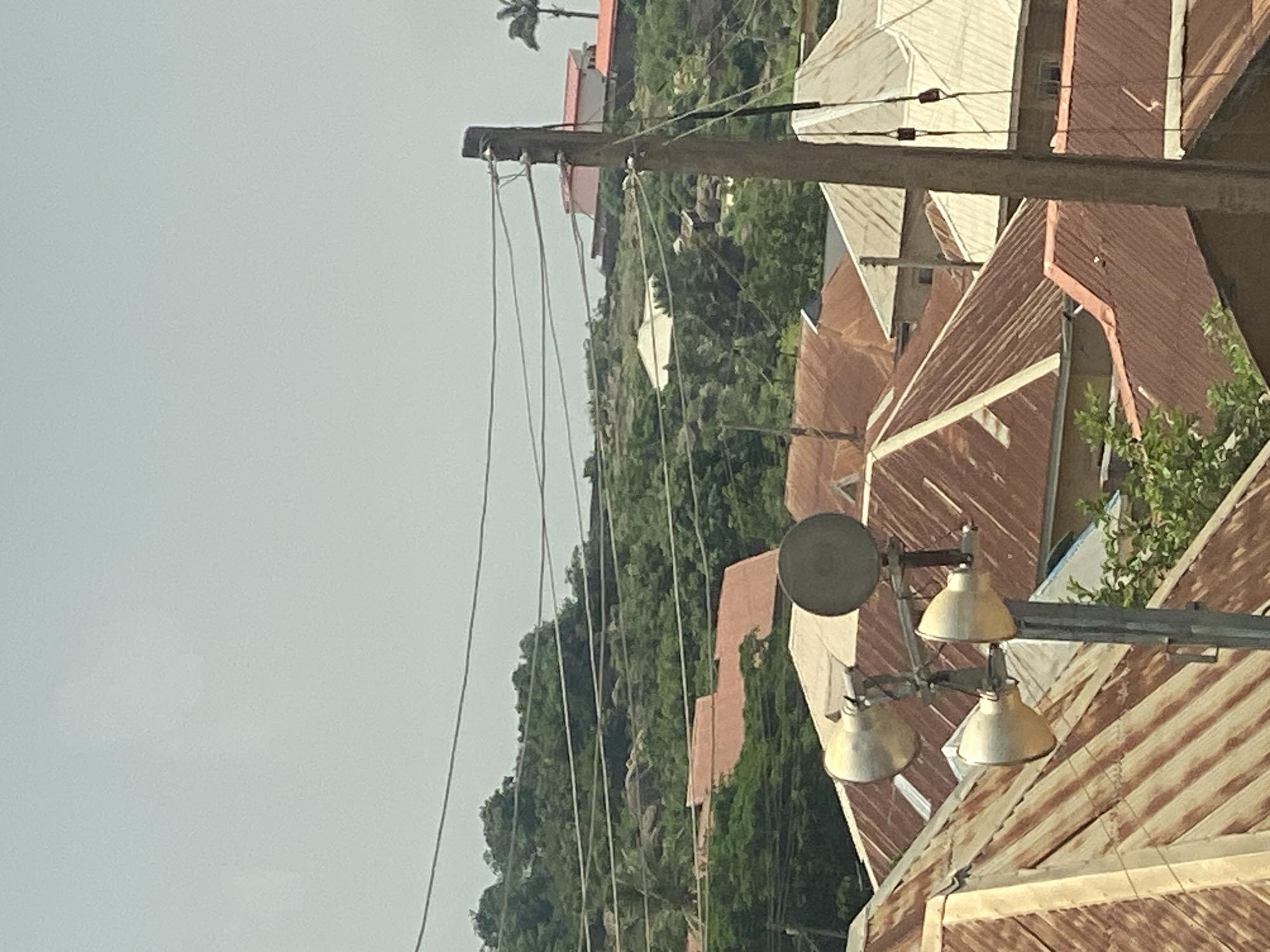 This image depicts a cluster of buildings with corrugated metal roofs, situated on a hillside covered in lush greenery. Power lines crisscross the foreground, supported by a wooden utility pole. Several lights, possibly streetlights, are mounted on a pole in the mid-ground, casting a warm glow on the surrounding structures. The architecture suggests a rural or semi-urban setting in Jos, Nigeria, characterized by its hilly terrain and common use of metal roofing. The sky is overcast, indicating a cloudy day, and the overall atmosphere appears serene. No people are visible in the frame, focusing the attention on the man-made structures and the natural landscape. The scene is likely captured during daylight hours, though the cloud cover diffuses the light. There is no visible text in the image.