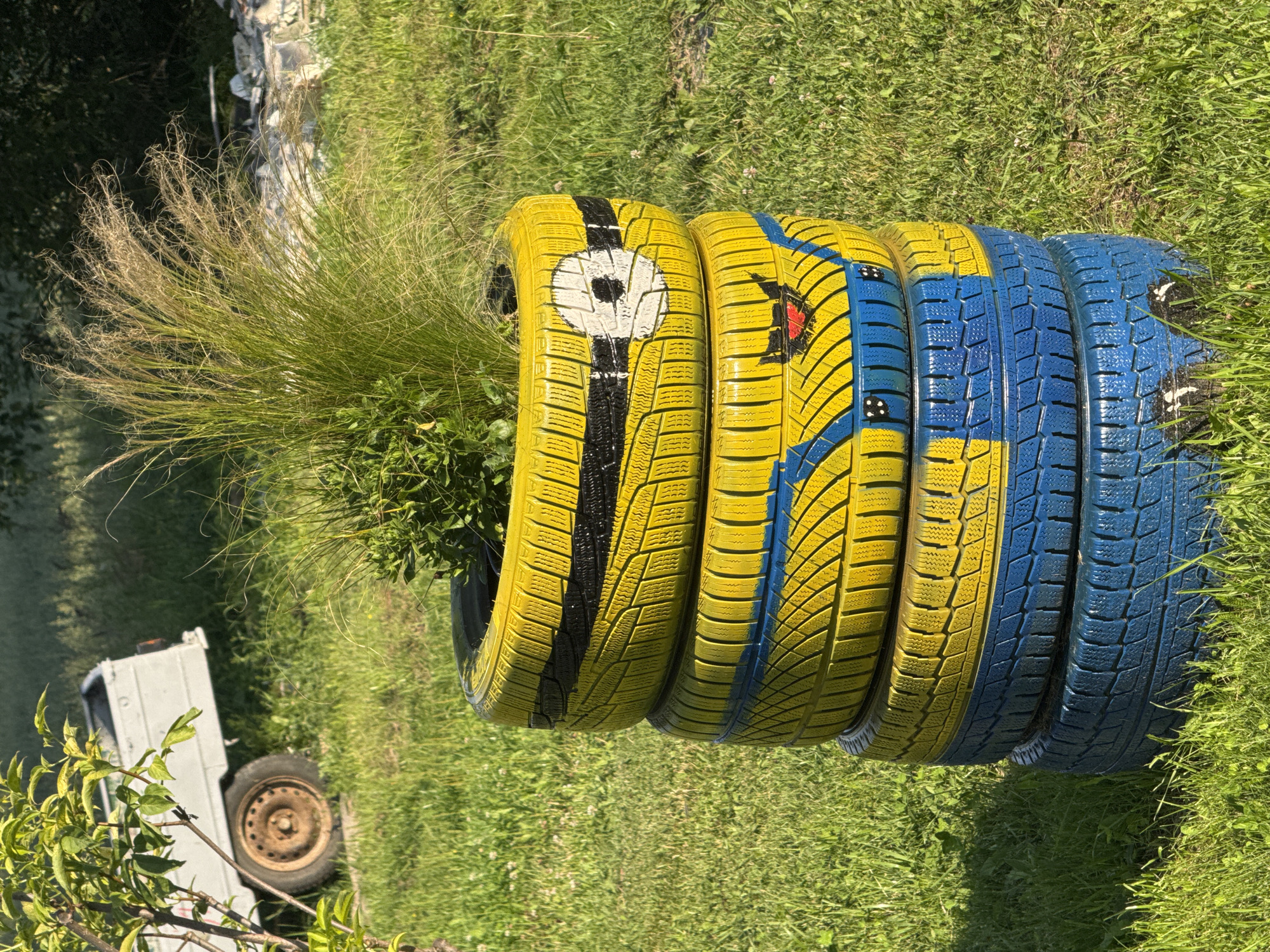 The image shows a stack of four painted tires serving as a planter in Pilskalns, Latvia. The tires are painted yellow and blue, resembling a Minion character. The top tire is yellow with a black stripe and a white circle with a black pupil, representing an eye. The second tire from the top is also yellow with blue accents and painted features, possibly a mouth. The third and fourth tires from the top are painted blue. A clump of tall, wispy grass and green shrubbery is planted in the top tire. The planter is situated on a grassy lawn, with a blurred background that includes trees and a white trailer with a rusty wheel visible on the left side of the image. The lighting suggests it is daytime, with sunlight casting shadows.