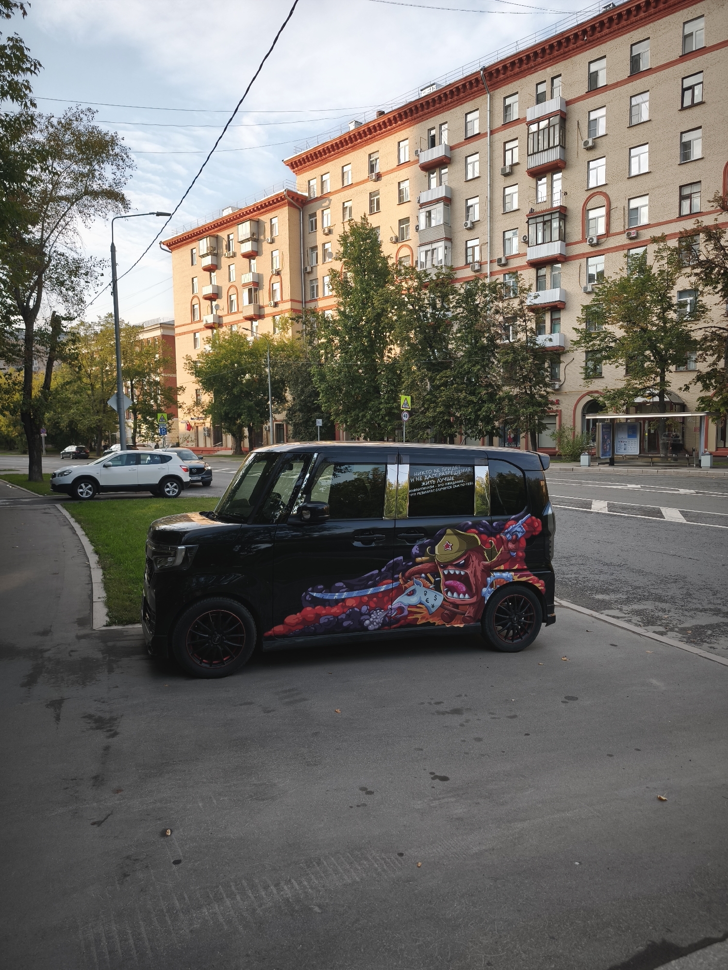 The image captures a street scene in Moscow, Russia, on a cloudy day. In the foreground, a black car with a cartoonish, vibrant mural on its side is parked on the side of the road. The mural features a monster-like character with a red hat and a pink body, surrounded by colorful abstract shapes and what appears to be money symbols. The car has red accents on its wheels.
To the left of the car, a paved sidewalk and a grassy area are visible, with a white SUV parked further down the street. Behind the car, a multi-story building with light-colored brick facade and red trim dominates the background. The building has numerous windows and balconies. Trees with green foliage line the street, and power lines are strung across the sky. A bus stop shelter is located across the street from the car. The overall environment suggests a residential or mixed-use urban area.