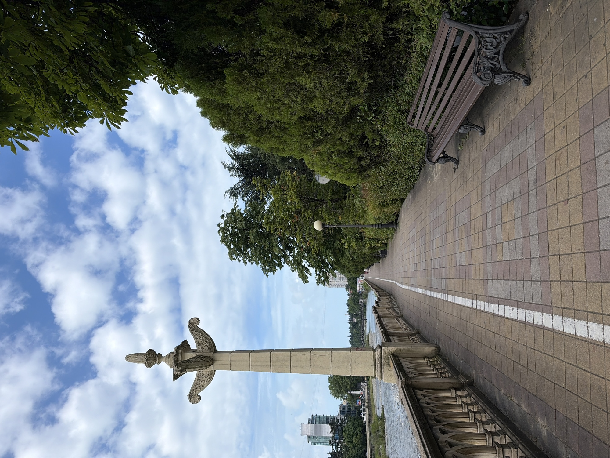 The image depicts a paved walkway along a river in Sochi, Russia. The walkway is lined with a decorative stone balustrade on the left, and a dense row of green bushes and trees on the right. A wooden park bench with ornate cast-iron legs sits on the right side of the walkway. In the distance, several buildings and bridges are visible, and a few people are walking along the path. The sky above is partly cloudy with a mix of blue sky and white clouds. A decorative stone pillar with an ornate top stands prominently on the left side of the walkway. The river water appears somewhat murky and flows gently.