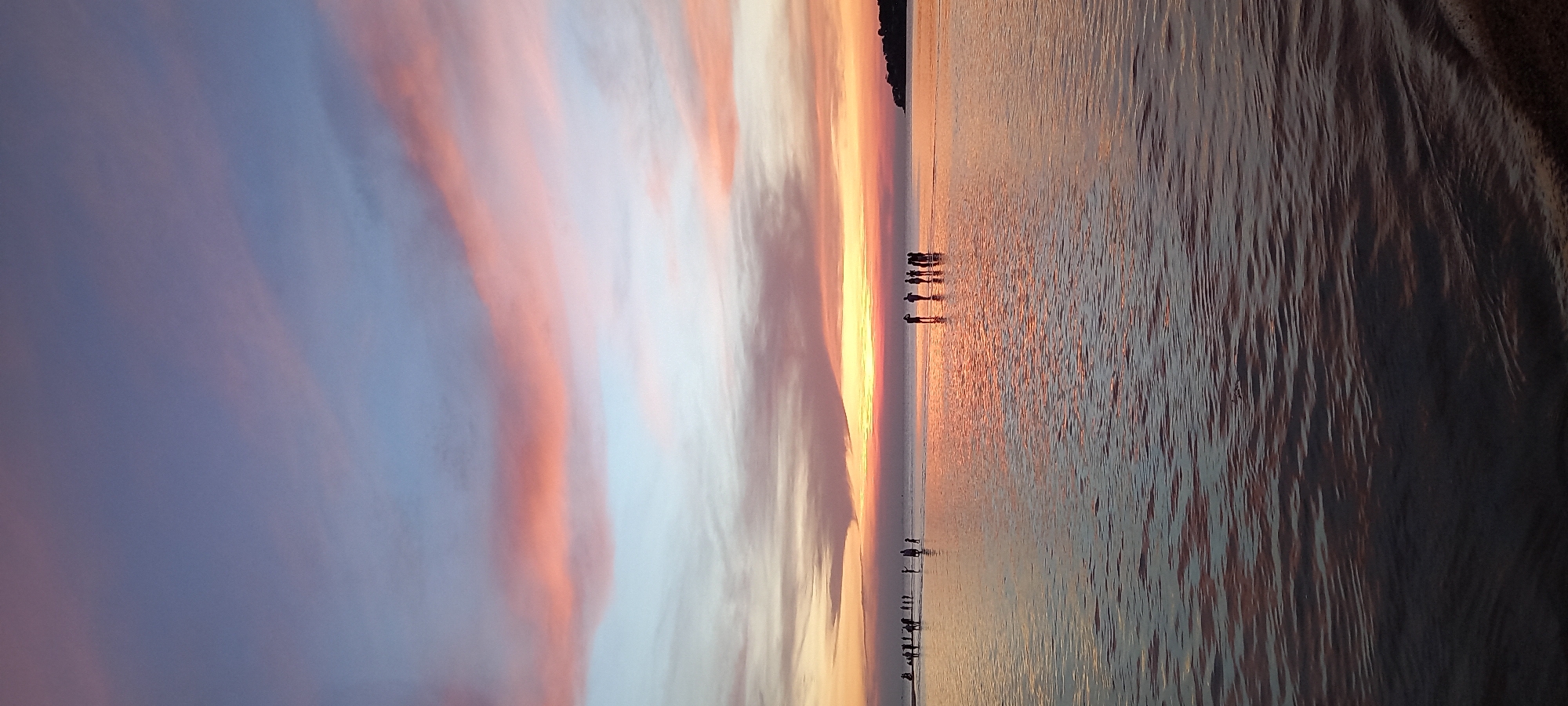 The image depicts a breathtaking sunset or sunrise over a calm body of water, likely a beach in Baan Sri Thanu, Thailand. The sky, occupying the upper left, is a canvas of soft blues, purples, and vibrant orange and pink hues, suggesting the transition of day to night or night to day. Wispy clouds are streaked across the horizon, illuminated by the low-lying sun.
The tranquil water surface, stretching across the middle and lower right, reflects the sky's vivid colors, creating a shimmering effect. A bright, warm glow emanates from the horizon where the sun is setting or rising, casting a luminous path across the water.
Several groups of people are visible as dark silhouettes against this radiant backdrop. They appear to be standing or walking in the shallow water, observing the natural spectacle. In the mid-ground, a distinct group of about five individuals is gathered, while other smaller figures are scattered further along the water's edge. Their indistinct forms emphasize the vastness of the scene and their contemplative presence within it.
A faint, dark landmass, possibly a distant island or shoreline, is visible on the far right edge of the horizon. The atmosphere conveyed is one of profound peace and natural beauty, with the dramatic colors of the sky and their reflection on the water being the central focus. There is no visible text in the image.
