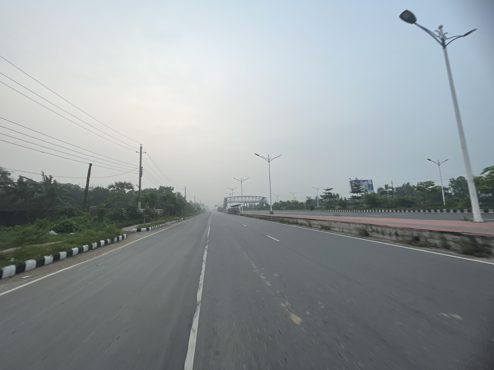 The image depicts a straight, multi-lane road in Purbachal, Bangladesh, likely a highway or major thoroughfare. The road surface is grey asphalt, with a white dividing line. There are sidewalks with black and white barriers and concrete median barrier with red section on either side of the road. Street lights line both sides of the road, with power lines visible on the left. In the distance, a pedestrian bridge crosses the road. Vegetation and trees are present along the roadside. The sky is overcast and hazy. No people are visible in the image.