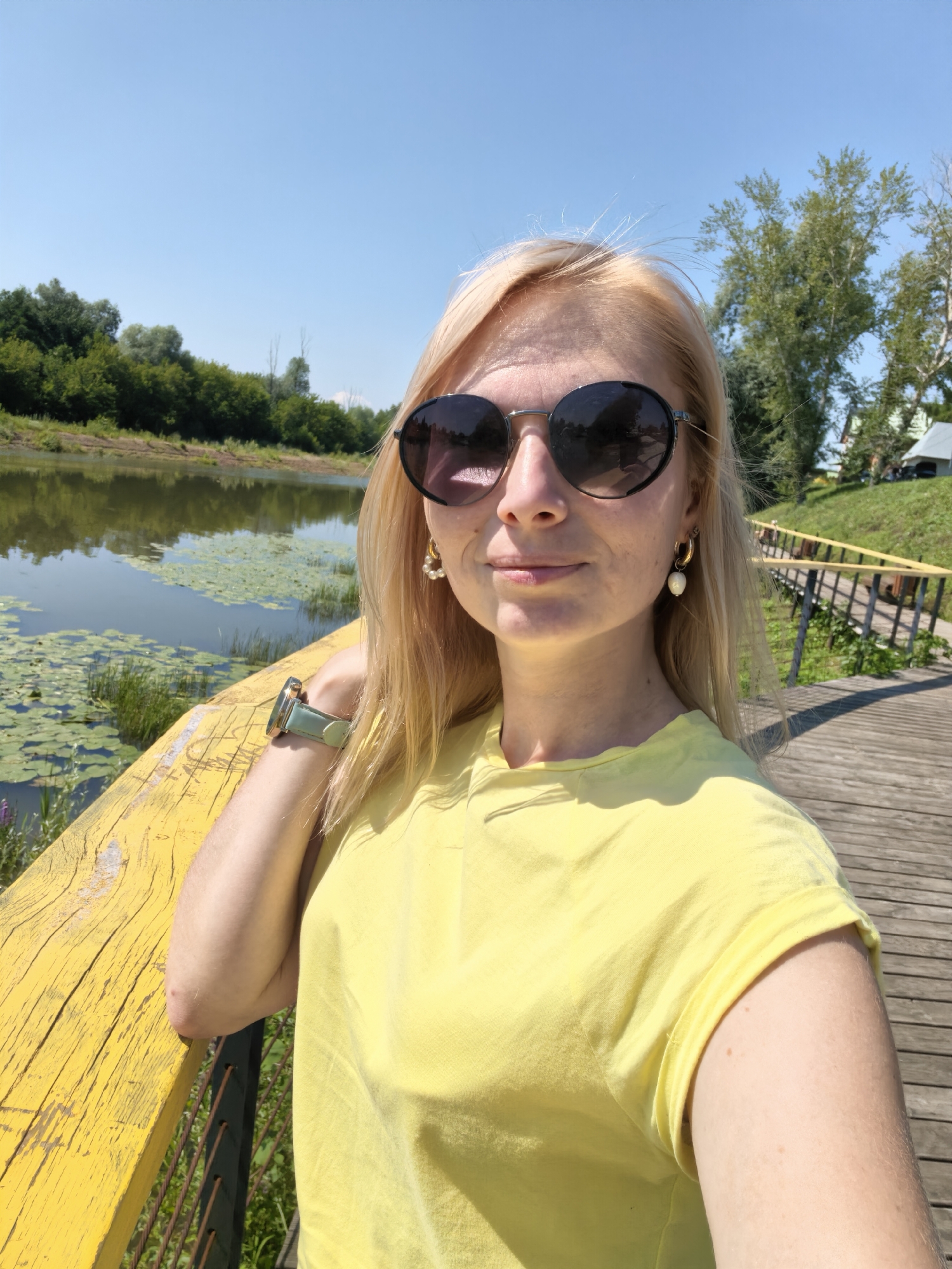 A blonde woman wearing sunglasses and a yellow t-shirt is taking a selfie on a wooden boardwalk next to a calm body of water. Lily pads are visible on the water's surface. The background shows a lush green treeline and a clear blue sky. The boardwalk has a bright yellow railing. The woman has earrings and a watch on her left wrist. The scene appears to be outdoors in a park or recreational area. There is no identifiable information to connect this image to Sterlitamak, Russia.