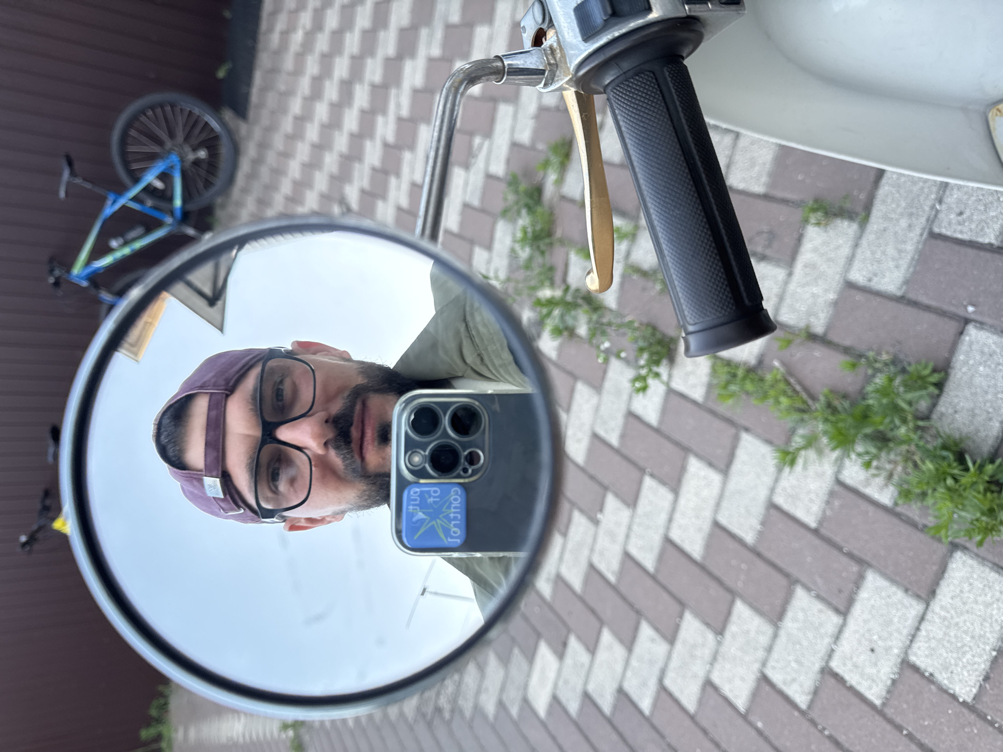 The image is a first-person perspective selfie taken in a mirror, likely of a motorcycle or scooter. The person in the mirror is wearing a purple baseball cap and glasses. They have a beard and are looking directly at the camera. They are holding a smartphone in their hand, which is also visible in the reflection, displaying a sticker with blue and yellow text that reads "CONTROL".
The background reflected in the mirror shows a blue and green bicycle with black wheels leaning against a brown fence. The ground is paved with interlocking bricks in a pattern of brown and light gray. There are patches of green foliage growing between the bricks. The sky appears overcast. The image seems to be taken outdoors, possibly in a residential area, given the fence and paved ground. The lighting suggests it is daytime. The location is identified as Novaya Usman', Russia.