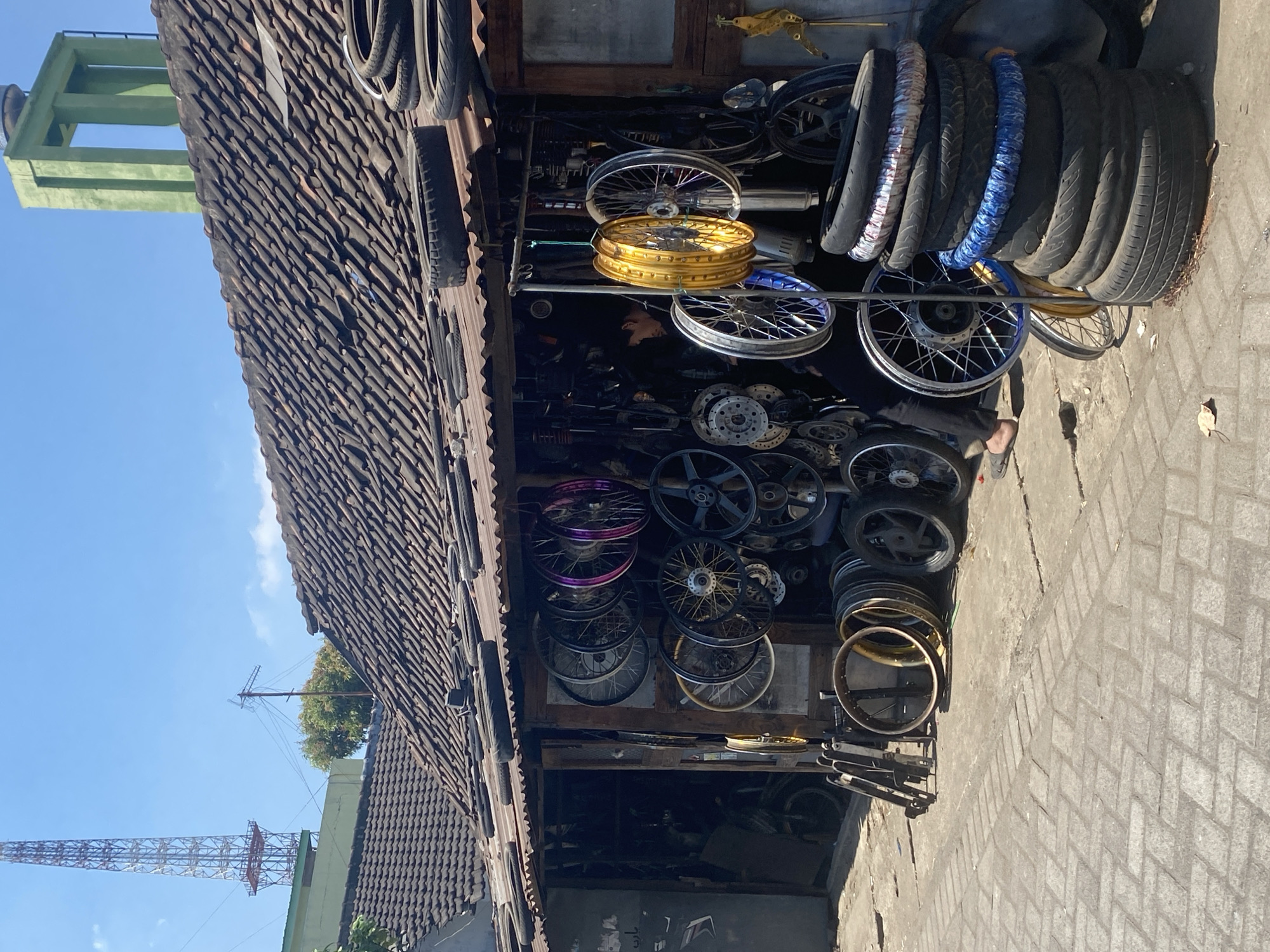 The image depicts an outdoor motorcycle parts shop located on a paved street in Kediri City, Indonesia. The shop features an open-air front with a corrugated metal roof supported by wooden beams. Various motorcycle wheels, some with colorful spokes, are displayed hanging from the eaves and on racks. Tires are stacked on the ground and on the roof of the shop. A person, wearing dark clothing and sandals, is visible inside the shop, appearing to be working. In the background, a tall radio tower and a green concrete structure with a water tank are prominent features of the city skyline. The scene is illuminated by bright sunlight, casting shadows on the ground.