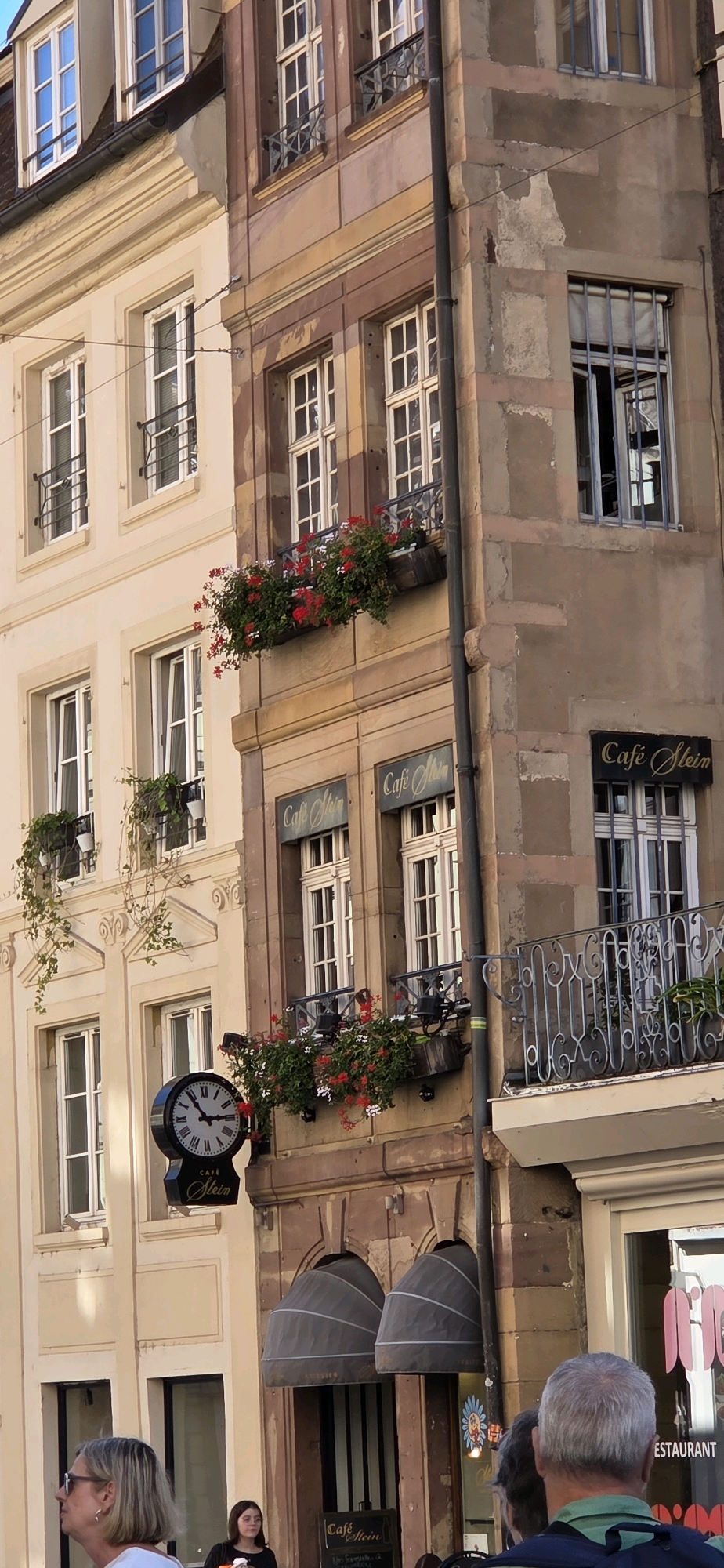 This image captures the exterior of a building in Strasbourg, France, likely located in the historic city center, as suggested by the classic European architecture. The building features multiple stories with distinct facades. The left side of the building is painted a light beige with classical architectural details, including ornate molding around the windows and decorative elements on the facade. The windows are tall and rectangular, with white frames and multiple panes, many featuring black wrought-iron grilles or small balconies.
The right side of the building presents a different facade, constructed with stone in shades of brown and pinkish-tan. This side is more weathered, with some peeling plaster revealing the underlying stonework. It features more windows, also with white frames, and some have wrought-iron bars. Several windows have planters with bright red and white flowers, adding a touch of color. Two signs with the text "Cafe Stein" are visible on this side, indicating the presence of a cafe. A round clock with Roman numerals hangs on the left side of the building, also with a "Cafe Stein" sign below it.
The environment appears to be a street scene. In the foreground, several people are visible. A woman with short blonde hair and sunglasses is looking to the left. Behind her, another woman with dark hair stands. Further to the right, a man with graying hair, facing away from the camera, is wearing a green shirt and a backpack. The lower part of the image shows the entrance to the cafe, with arched awnings and striped vertical elements inside. A part of a sign with the word "RESTAURANT" is visible on the far right. The overall impression is of a charming European street with a lively cafe.