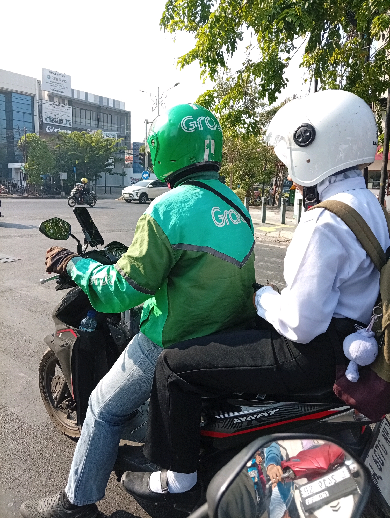 Two people are on a motorcycle in Surabaya, Indonesia. The driver wears a green helmet and jacket with "Grab" logo. The passenger wears a white helmet and a white shirt with a dark backpack. They are stopped at an intersection with a white car approaching. A building with a large advertisement is visible in the background. The motorcycle is a black and red Honda Beat. The scene is captured from a low angle, likely from another motorcycle.
