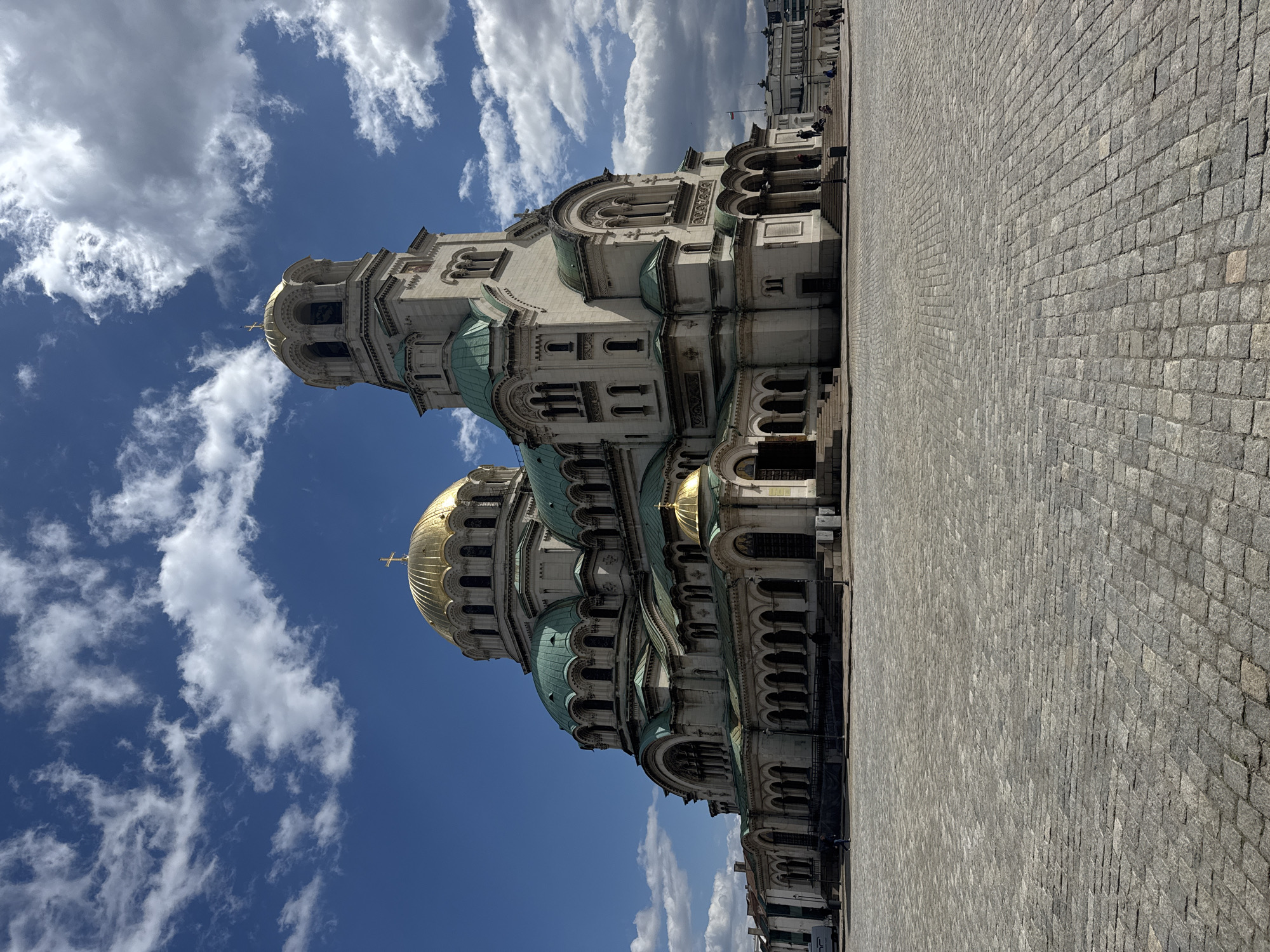 The image shows the Alexander Nevsky Cathedral in Sofia, Bulgaria, from a low angle, emphasizing its scale. The cathedral is white with gold domes and green accents, set against a bright blue sky with scattered white clouds. In the background, the cobblestone pavement stretches into the distance, occupying a large portion of the frame. People can be seen standing near the edge of the pavement, close to other buildings. The scene appears to be captured on a sunny day, with clear visibility.