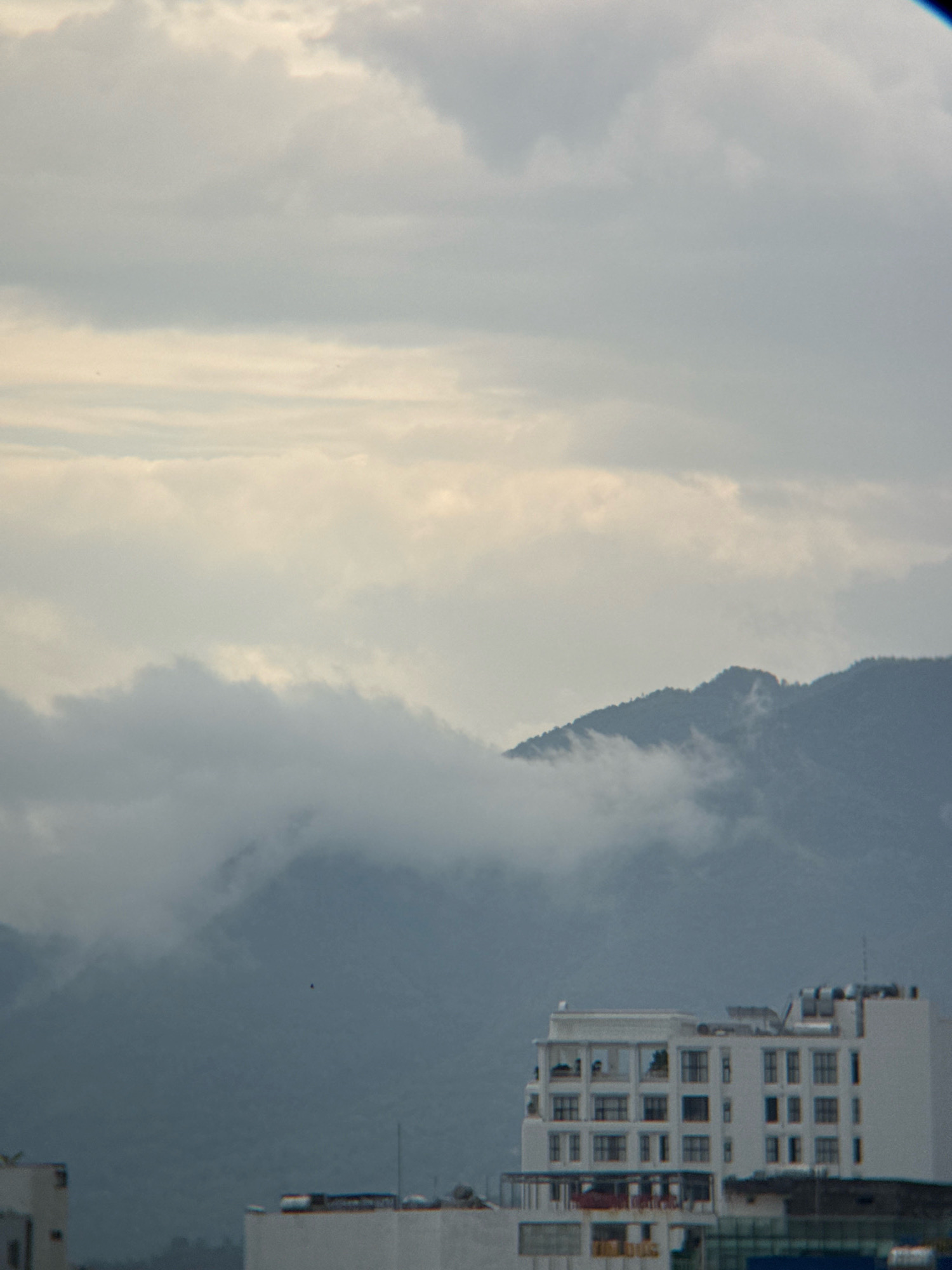 The image depicts a hazy, overcast sky above the city of Nha Trang, Vietnam. In the foreground, a modern white building with multiple floors and many windows is visible, with a rooftop featuring solar panels and other equipment. Partially obscured by clouds in the middle ground is a lush, green mountainside. The overall atmosphere is muted, with soft lighting due to the cloudy conditions.