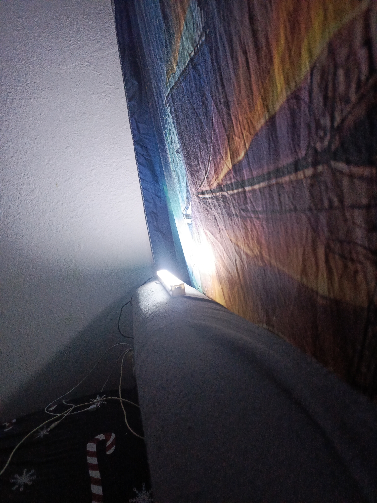 A bright LED light strip is positioned on a grey upholstered surface, casting light upwards onto a textured white wall and a draped tapestry. The tapestry, with its creased fabric, displays a pattern of abstract, fiery colors ranging from deep blues and purples to oranges and yellows. Below the light strip, on a dark surface adorned with snowflake and candy cane motifs, are several white and yellow electrical cords. The scene appears to be indoors, possibly a domestic setting, with no indication of Bolesławiec, Poland.