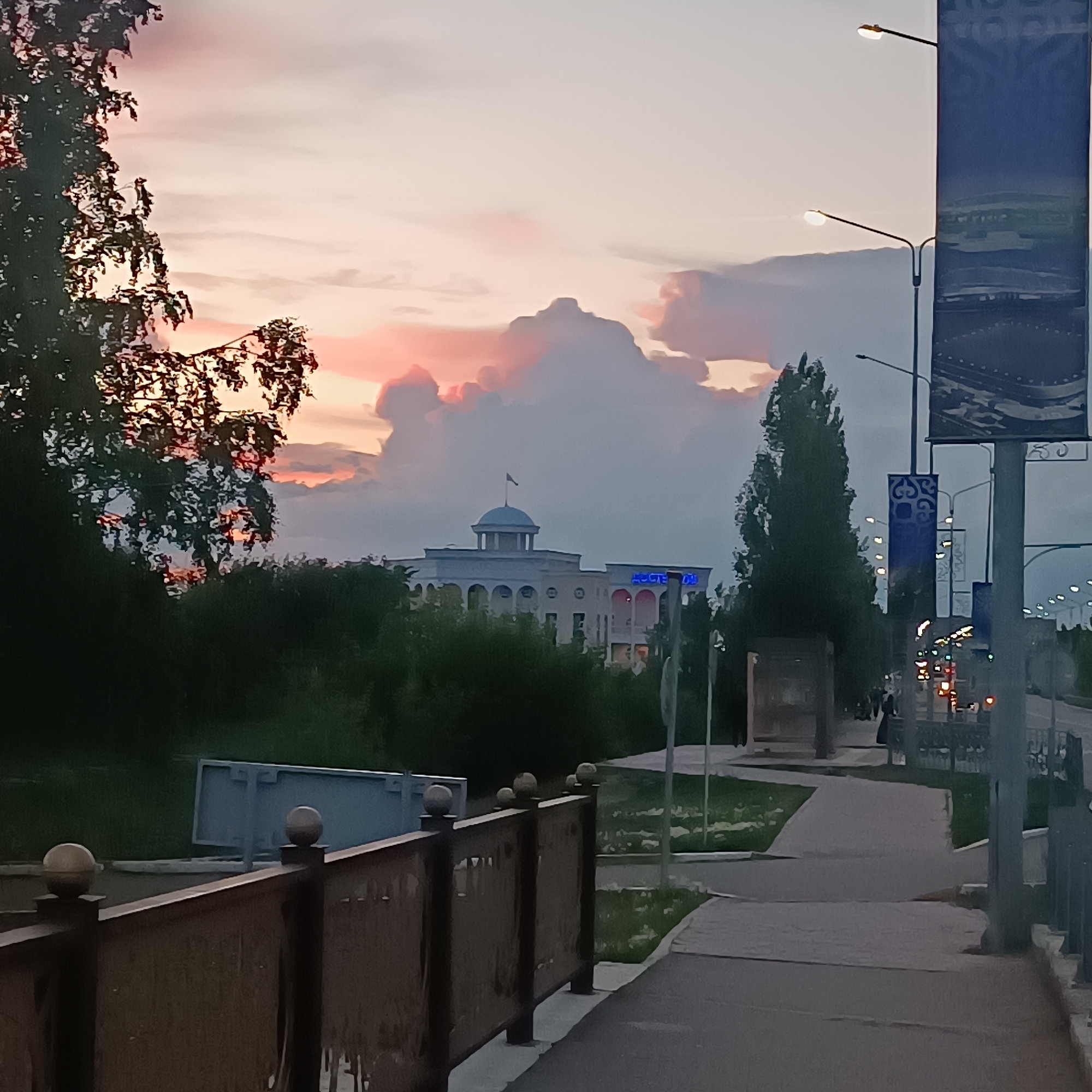 The image depicts a street scene in Kokshetau, Kazakhstan, during dusk. A large, light-colored building with arched windows and a dome is visible in the background, partially obscured by trees. The building has a flag on top and neon blue lettering that reads "HOSTELL" above an entrance. The sky is filled with dramatic clouds, illuminated with pink and orange hues from the setting sun.
In the foreground, a dark wooden fence with decorative spheres lines a sidewalk. Across the street, there are streetlights and a large vertical banner with intricate blue patterns. The street is paved, and there are grassy areas bordering the sidewalk. A few streetlights are on, casting a warm glow. The overall atmosphere is calm and serene, with the vibrant sky contrasting with the muted tones of the urban landscape. There are no distinct individuals clearly visible in the frame, suggesting a quiet moment in the city.