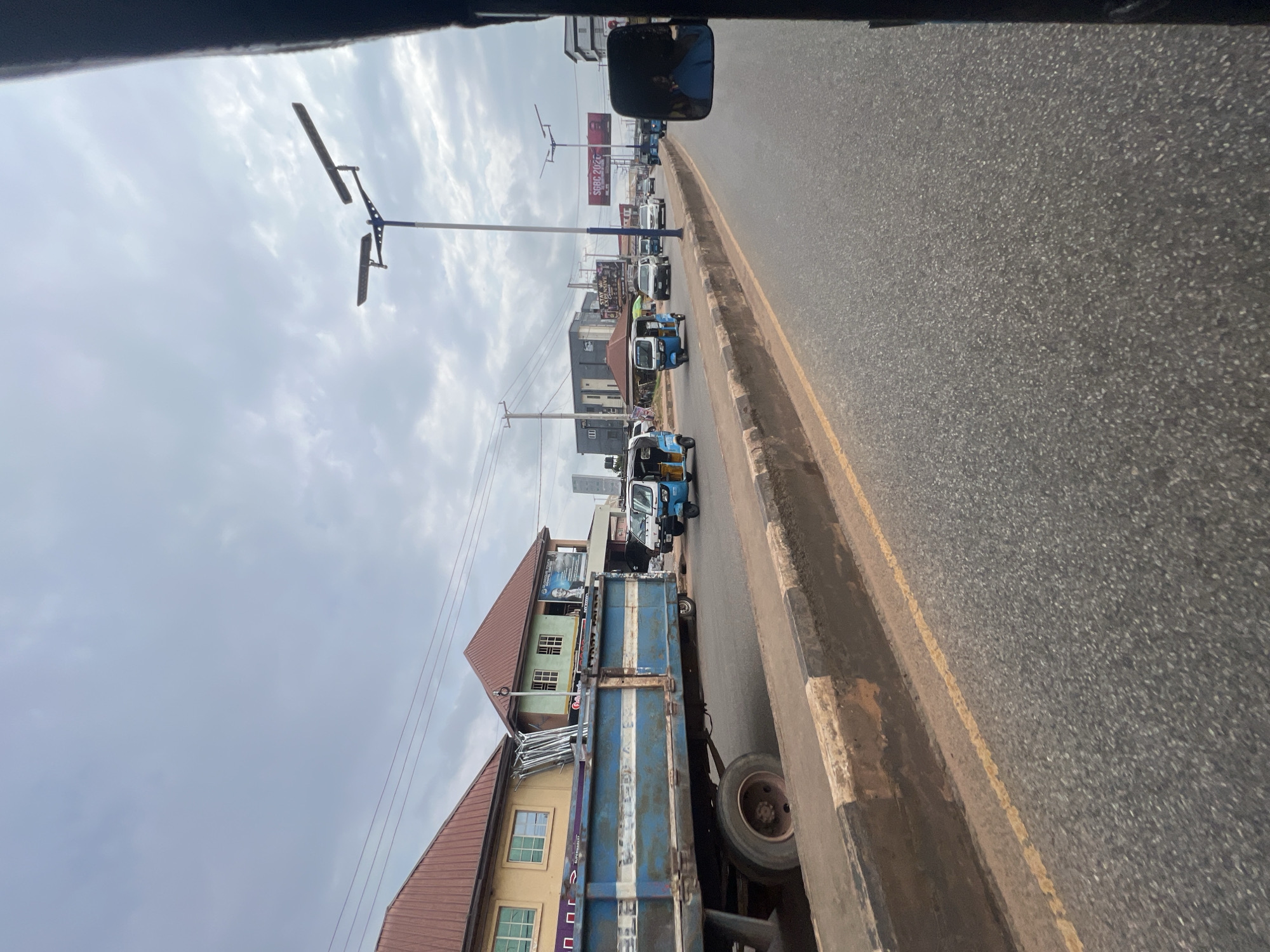 The image depicts a street scene in Okpanam, Nigeria. A blue and white truck with a flatbed is parked on the left side of the road, laden with what appears to be metal scaffolding. In front of the truck, several blue and white auto-rickshaws, commonly known as 'keke' or 'marwa', are in motion on the asphalt road. The road has a concrete barrier with black and white markings separating it from a sidewalk or median strip. Beyond the road, buildings with reddish-brown roofs are visible. The sky is overcast with grey clouds. A street light with two lamps stands tall on the right side of the road. A large billboard with text and images is also visible in the distance. The perspective suggests the image was taken from inside a moving vehicle, as indicated by the dark frame on the right edge of the photo, possibly a car door or window frame.