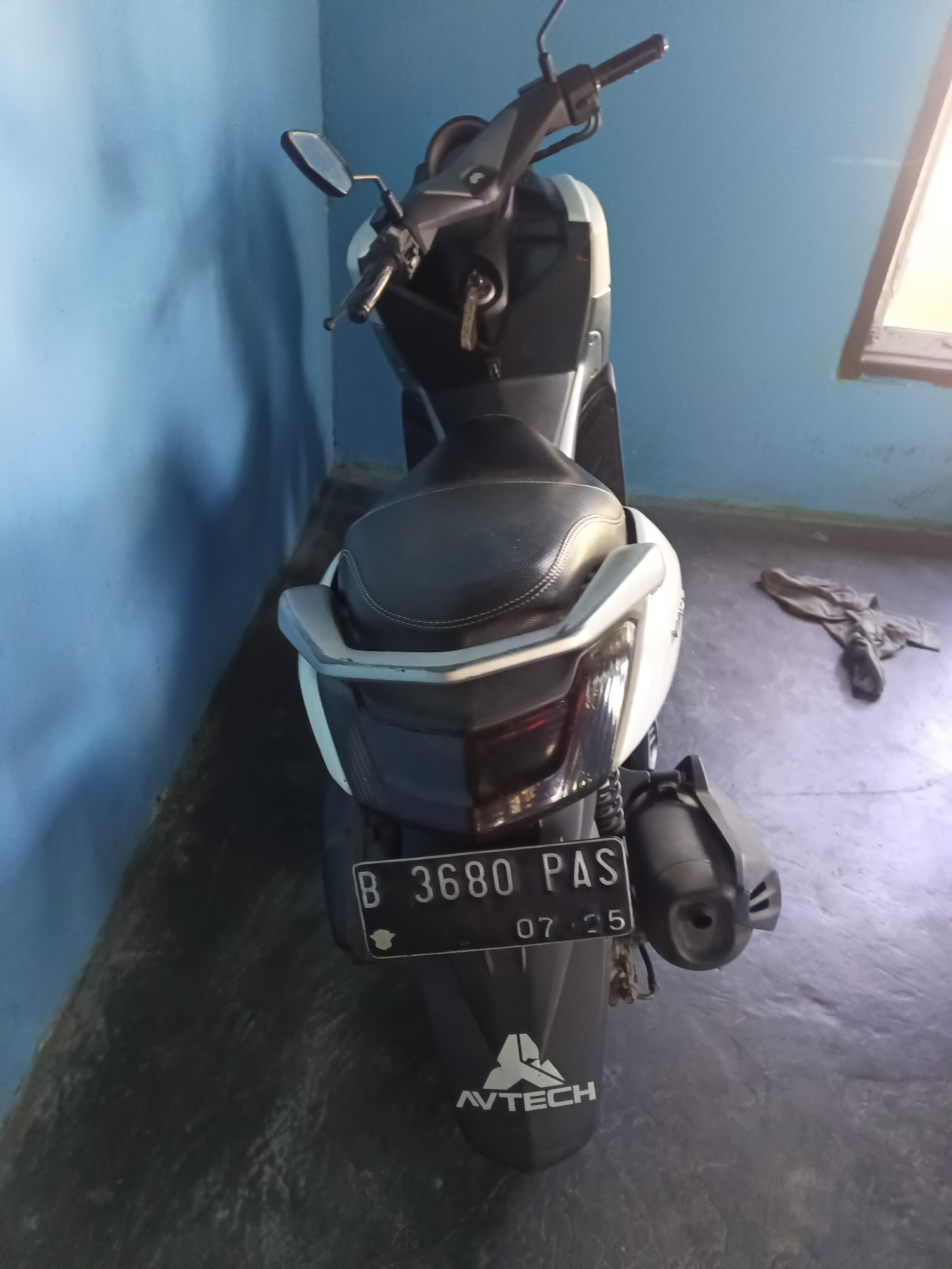 A rear view of a white and black scooter is captured in the city of Negara Nabung, Indonesia. The scooter is parked indoors, leaning against a vibrant blue wall. The license plate, clearly visible, reads "B 3680 PAS 07.25." A grey cloth is draped on the floor to the right of the scooter. A window with bright sunlight is visible in the background, casting a warm glow on the scene. The scooter's rear fender features a white logo with the text "AVTECH."