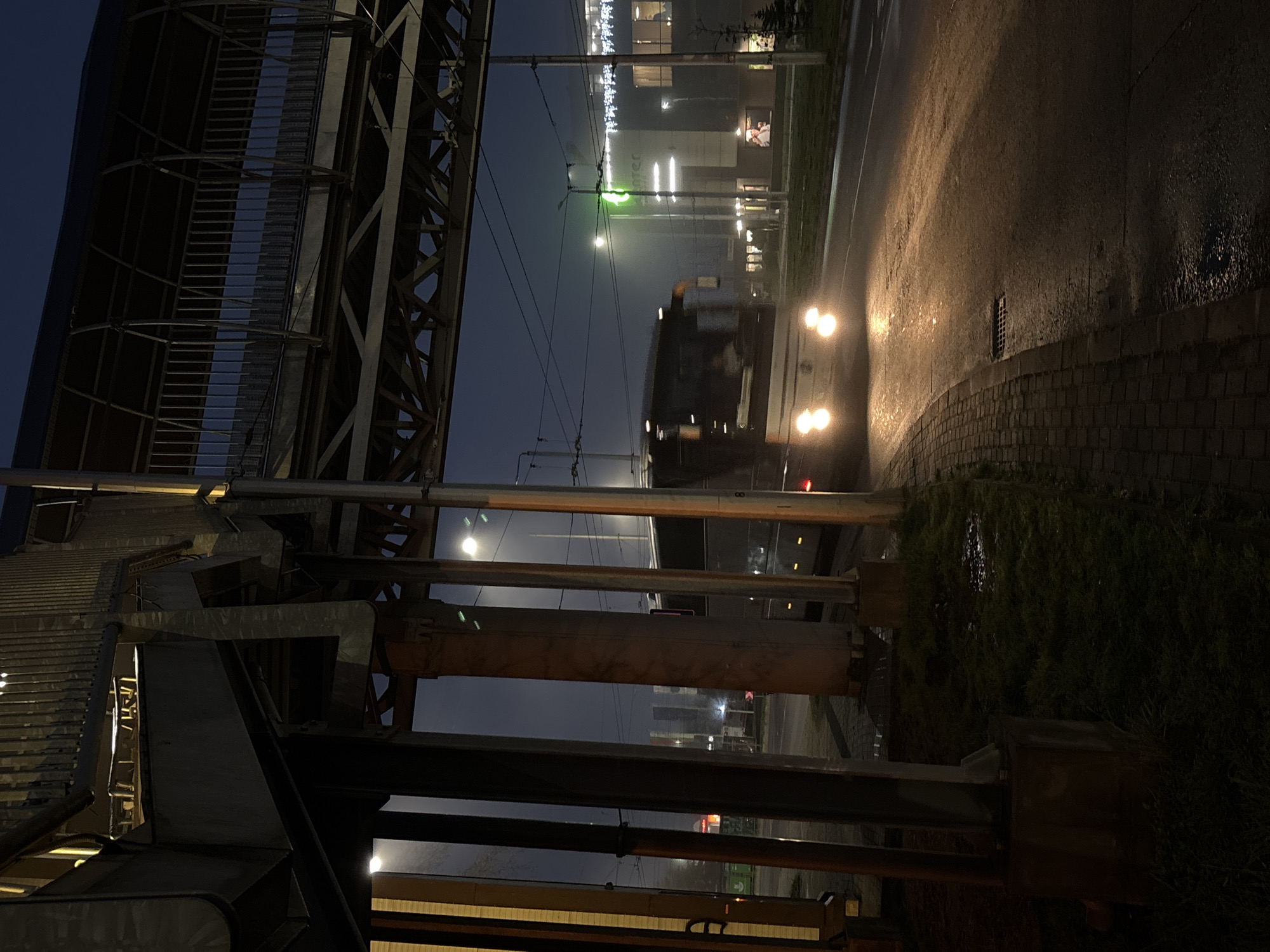 At night in Varna, Bulgaria, a bus moves on a wet road, its headlights reflecting off the slick surface. The view is partially obstructed by an elevated pedestrian walkway constructed of steel beams and supports. In the background, buildings with illuminated windows and streetlights are visible. The sky is dark, suggesting a late hour. Vegetation lines the roadside.