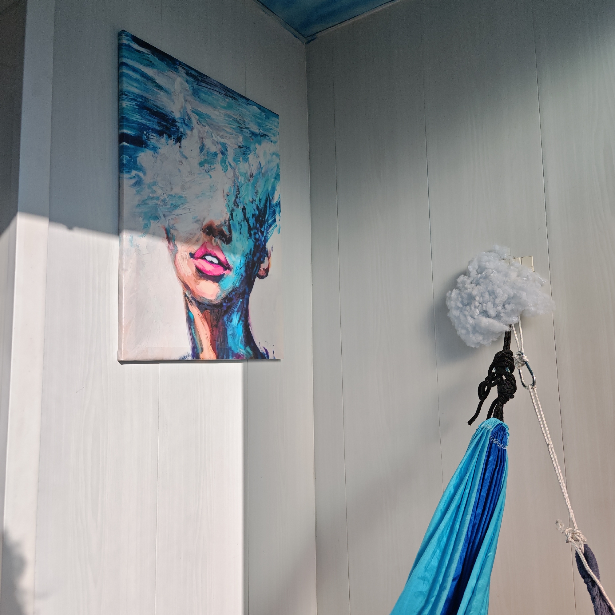 A corner of a room with white wooden paneling is visible. On the left wall hangs a painting of a person with their face obscured by an abstract explosion of blues and whites, but their pink lips are clearly visible. On the right wall, a white, fluffy, cloud-like object is hanging from a white plastic hook. Below that is suspended a folded blue hammock with black and white ropes. The ceiling is painted a light blue. The image seems to be taken in a well-lit space, likely indoors. There's no information indicating the location is Rostov, Russia.