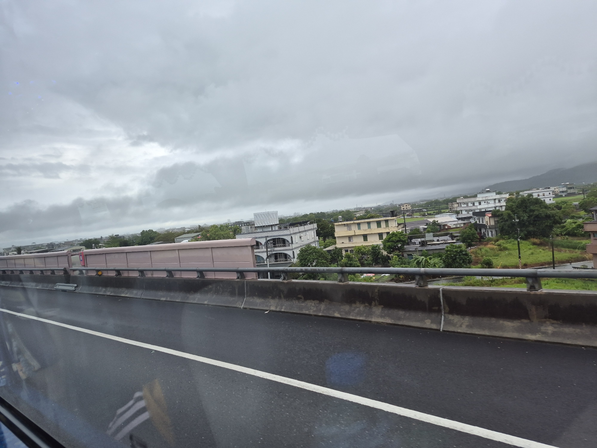 The image shows a view from a highway overpass in Jiaoxi Township, Taiwan, looking towards a residential area and the surrounding landscape under a cloudy sky. In the foreground, there is a wet asphalt road with a white line marking the lane. A concrete barrier with a metal railing separates the road from a pinkish-tan sound barrier wall. Beyond the sound barrier, buildings of various sizes and architectural styles are visible, interspersed with lush green trees and fields. The buildings appear to be a mix of residential and commercial structures, with some featuring multiple stories and balconies. In the background, rolling hills and mountains are faintly visible through the haze. The sky is heavily overcast, suggesting a cloudy or possibly rainy day. There are no people or vehicles clearly visible in the scene.
