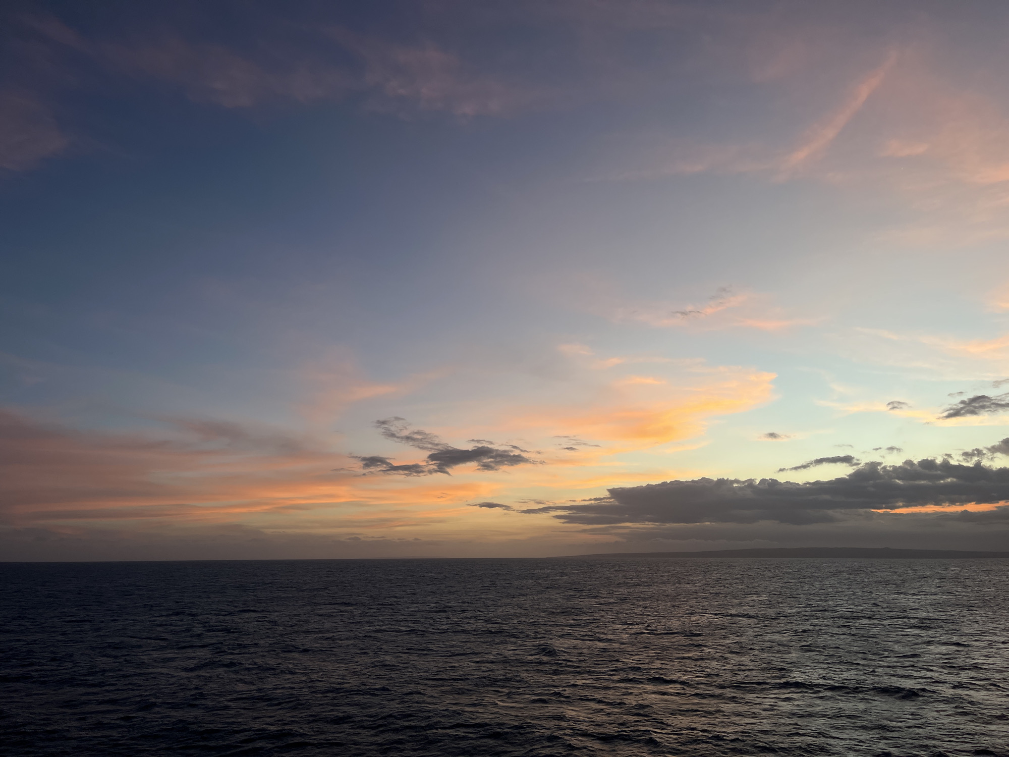 The image depicts a twilight scene over the ocean. The sky is filled with a mix of colors, transitioning from a deep blue at the top to pastel pinks, oranges, and yellows near the horizon. Clouds of varying sizes and shapes are scattered across the sky, some illuminated by the setting sun.
Below, the ocean is dark and textured with gentle waves. A distant landmass is faintly visible on the horizon.