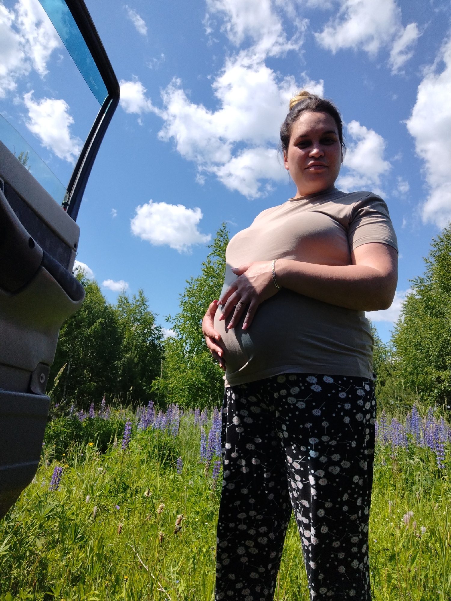 A pregnant woman stands in a field of grass and purple lupine flowers. She is wearing a beige t-shirt and black pants with a white floral pattern. The woman's hands are resting on her pregnant belly. To the left of the woman, a car door is open, revealing the interior of the vehicle. The background consists of green trees and a blue sky with white clouds. The scene appears to be set in a rural area, possibly in or near Zipunovo, Russia, given the presence of lupine flowers which are common in the region during summer.