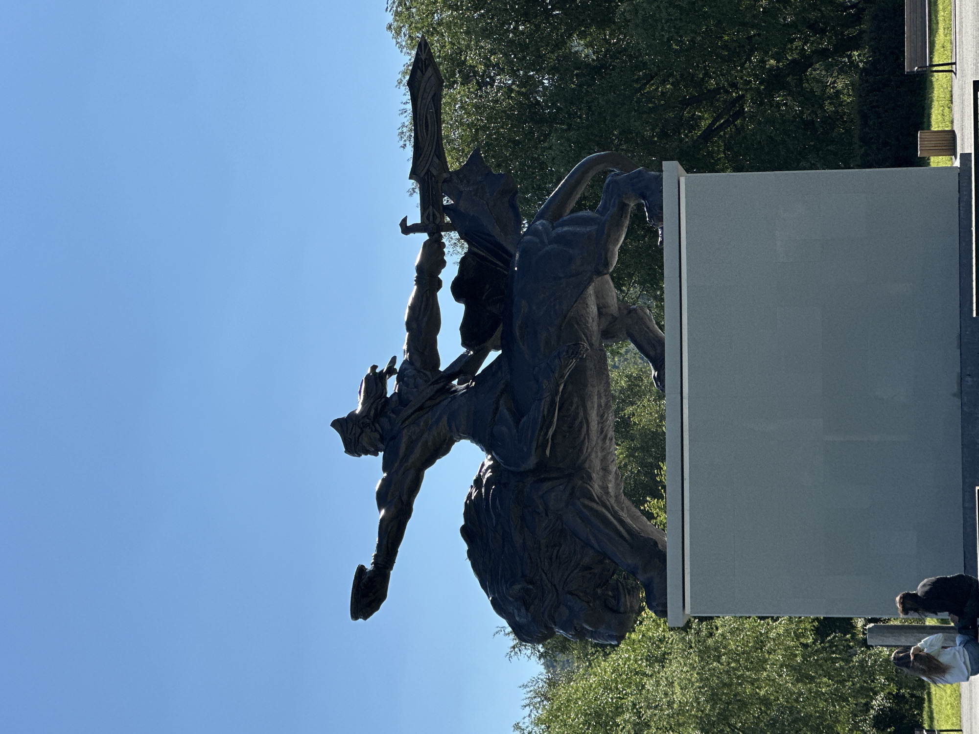 A dark, weathered bronze statue of a warrior riding a lion is displayed against a clear blue sky. The warrior, wearing a helmet and cloak, holds a sword aloft in his right hand and a bowl in his left. The lion, depicted in a powerful stride, has its mouth open. The statue stands on a large, light gray rectangular pedestal. In the foreground, at the base of the pedestal, two individuals are seated. The environment includes lush green trees and foliage in the background, with a glimpse of a park bench and trash receptacle on the right side. The overall scene is outdoors, bathed in sunlight.