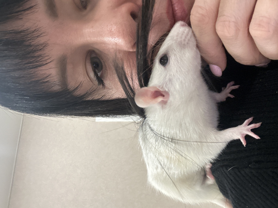 A person with dark hair and light eyes poses closely with a white rat. The rat is held near the person's face, its pink ears and tiny paws visible. The person's skin is fair and their fingernails are painted pale pink. The rat is perched on dark clothing. The background is a plain, light color. The image is oriented vertically with the person's face angled.