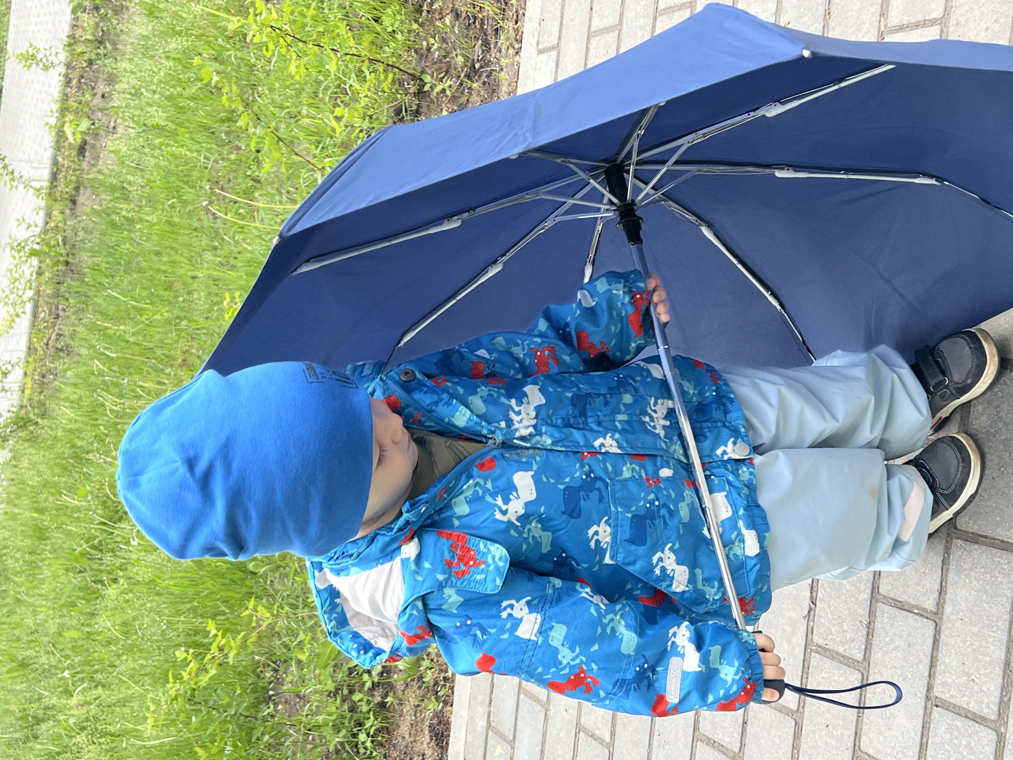 A child stands on a grey brick sidewalk under a blue umbrella, their body oriented towards the right side of the frame. The child wears a blue beanie, a blue jacket with red and white cartoon-like figures, light blue pants, and dark sneakers. Green grass grows on the left side of the frame. Based on the jacket's logo, this image was taken in the city of Chaikovsky, Russia.