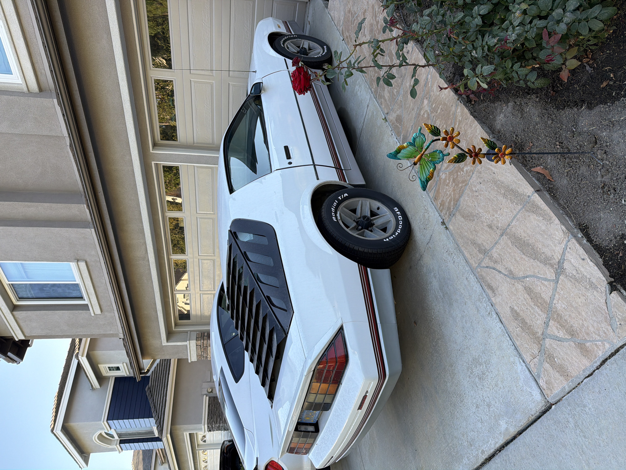 The image shows a white 1980s Chevrolet Camaro parked in the driveway of a two-story house in Aliso Viejo, California. The car is positioned to the right of a newer white Ford Mustang. The house features beige siding, beige garage doors, and brown tile roofing. A section of the house to the left is clad in blue horizontal siding with a large arched window. The driveway is paved with concrete and accented with stone pavers. In the foreground, a garden decoration of a butterfly and flowers is visible next to a rose bush. The sky is clear and blue.