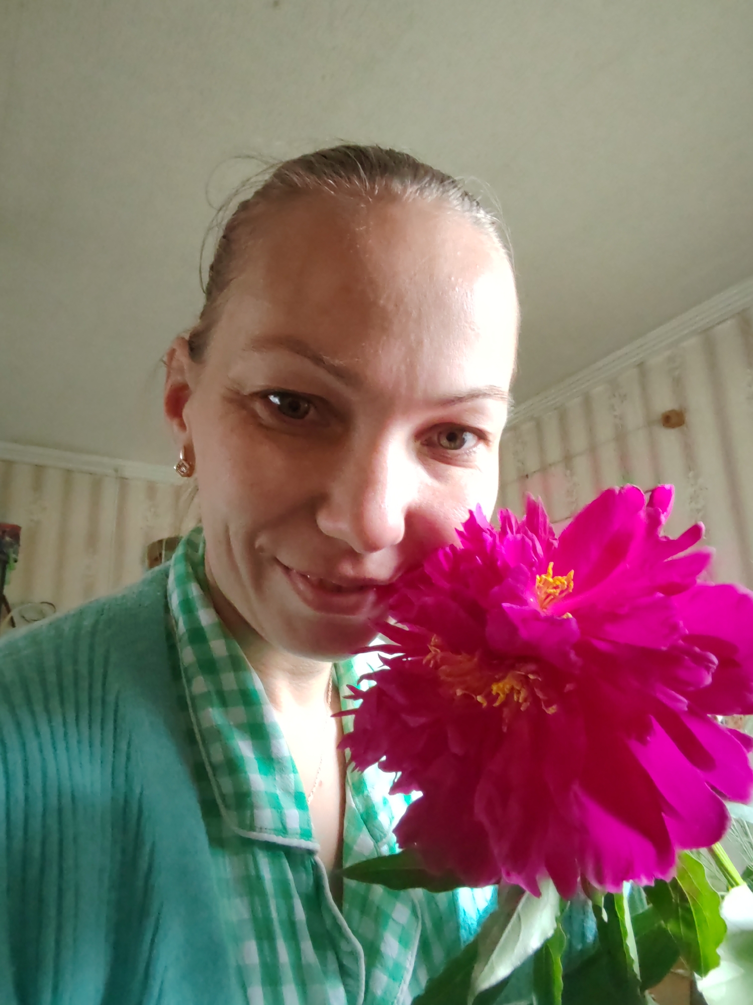 A woman is taking a selfie with a bright pink peony. She is wearing a light teal robe over a green and white checkered shirt. She has light brown hair pulled back from her face and is wearing a small hoop earring in her left ear. The background shows a wall with vertical striped wallpaper and a textured ceiling. The lighting suggests the photo was taken indoors during the day. The overall impression is a casual, personal moment. The location in the city of Чухлэм, Russia cannot be determined from the image itself.
