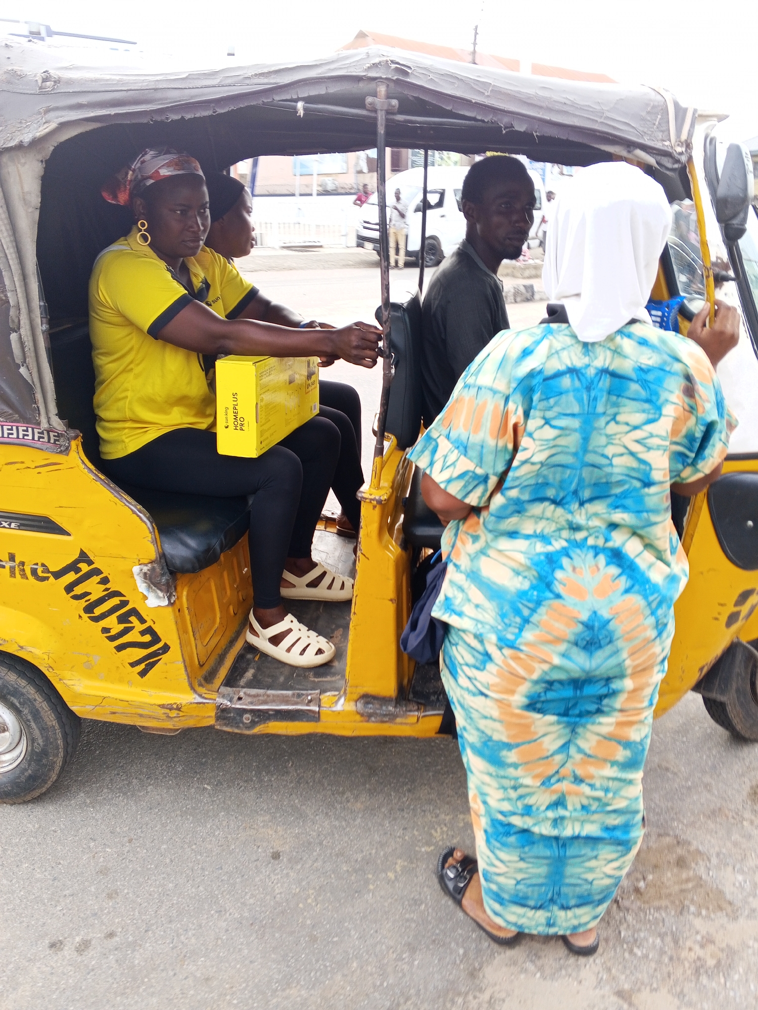 In Zango-ndaji, Nigeria, a yellow tricycle, possibly a "keke," is parked on an asphalt road. A woman wearing a yellow shirt, black leggings, and light-colored sandals sits inside, holding a yellow box labeled "HomePlus PRO." Another woman sits next to her. Behind them, a man is seated. Standing beside the tricycle, facing away from the camera, is a woman in a long, blue and beige tie-dye outfit and a white head covering. A few other people are visible in the background.